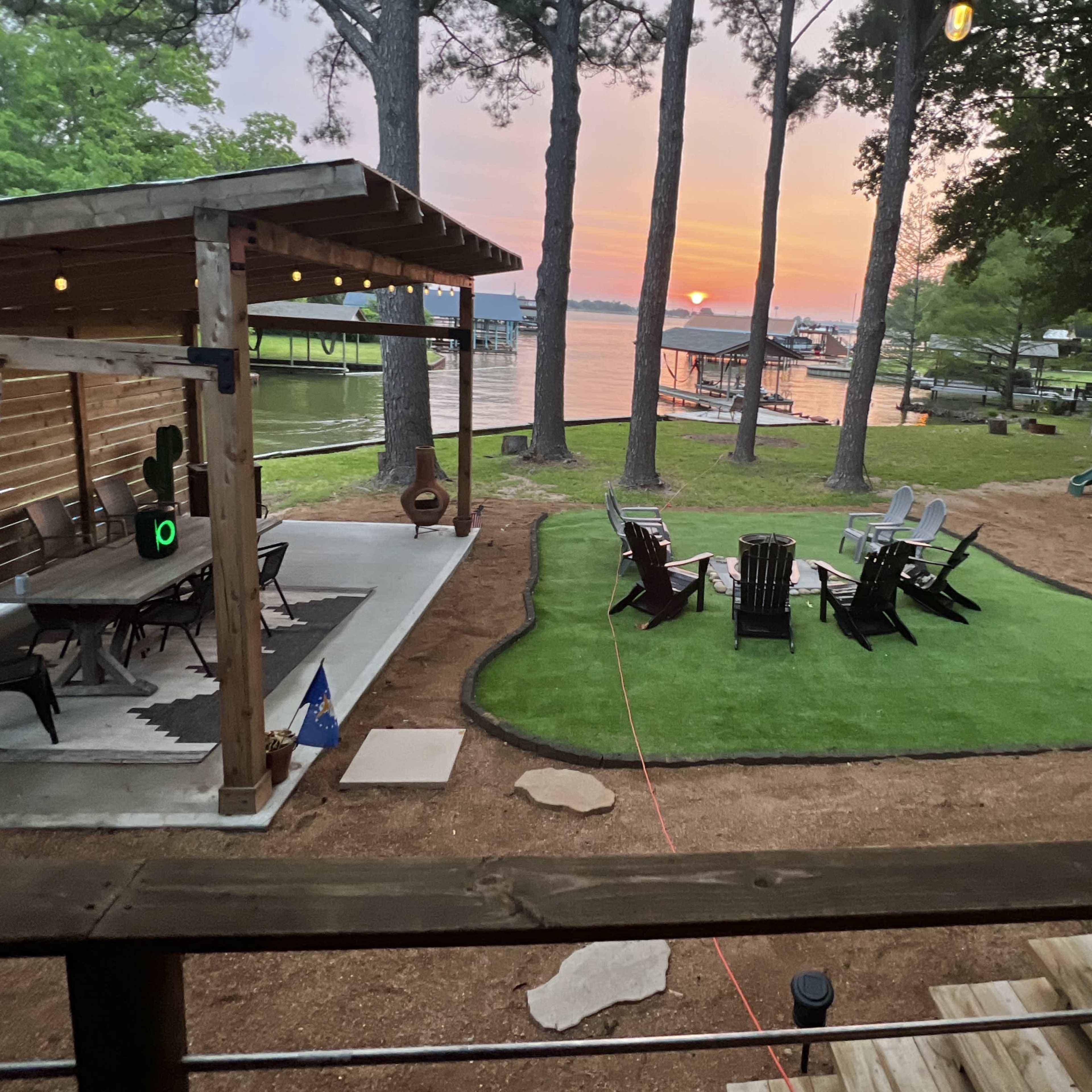 A lakeside setting with a covered patio, multiple lawn chairs arranged on a grassy area, and a view of the sunset reflecting on the water.