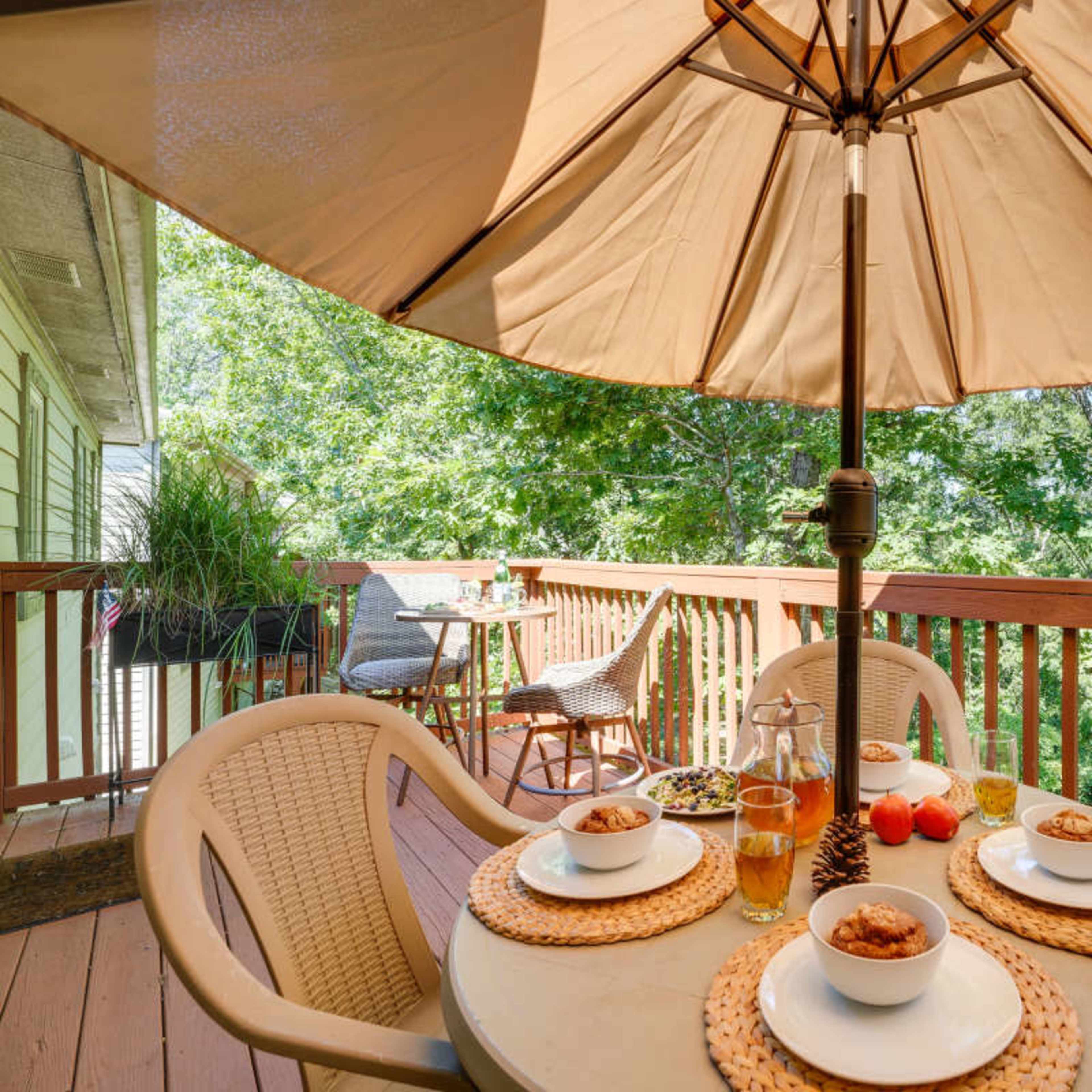 A table set with bowls of food and drinks is placed under a large umbrella on a deck overlooking greenery.