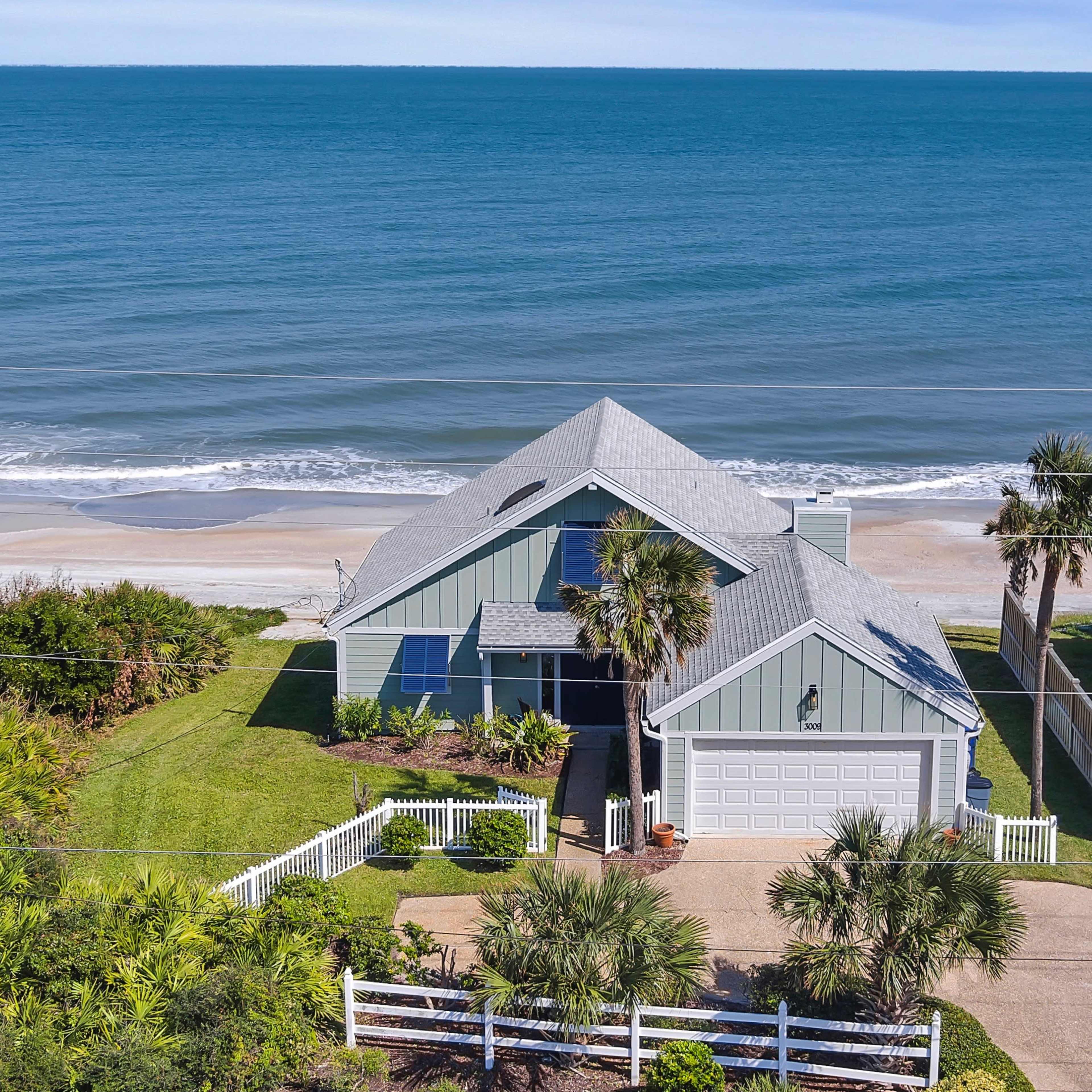 A coastal house with a gray roof and a beach visible in the background, surrounded by greenery and a white picket fence.