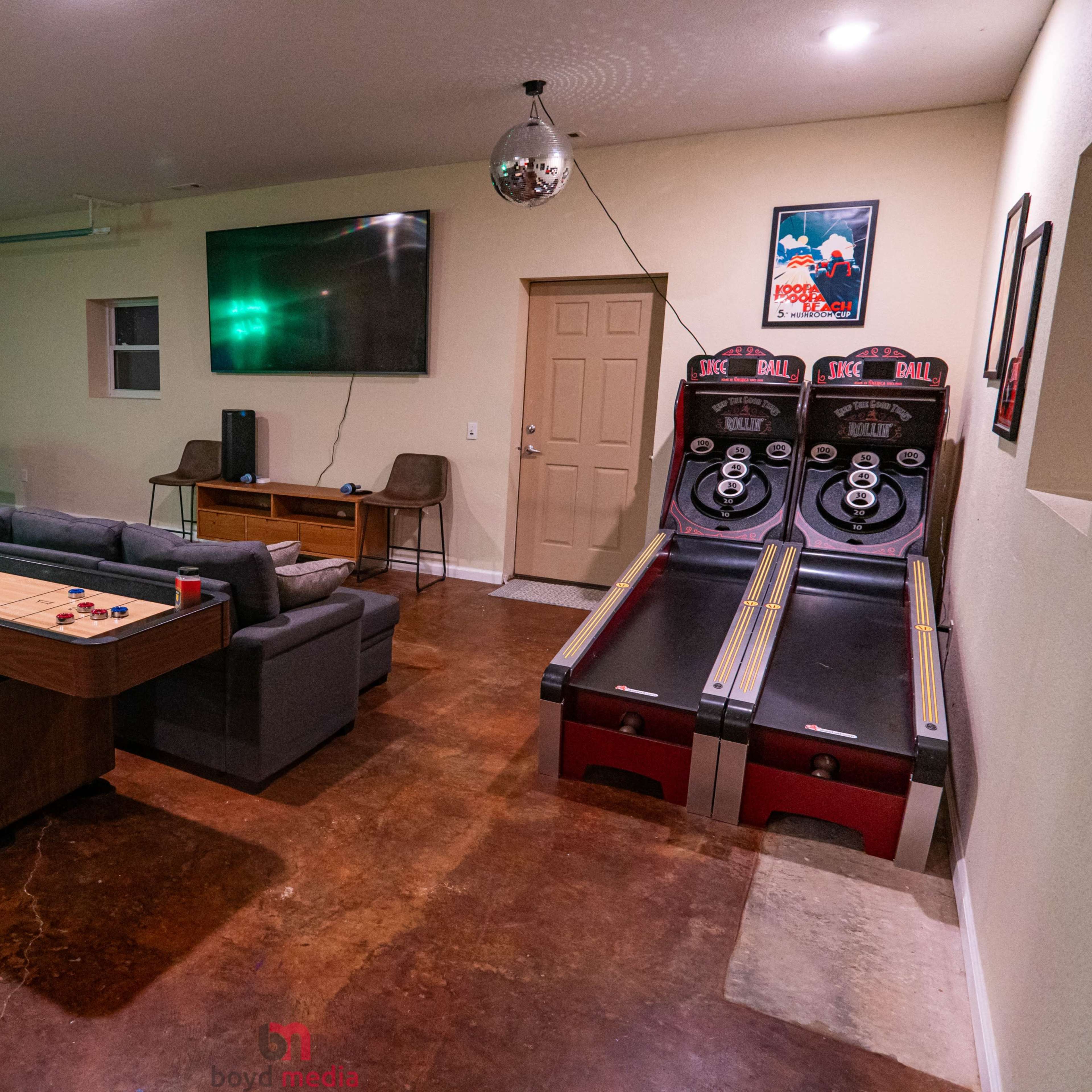 A recreational room with a couch, a television mounted on the wall, and two arcade-style skee-ball machines alongside a long wooden table.