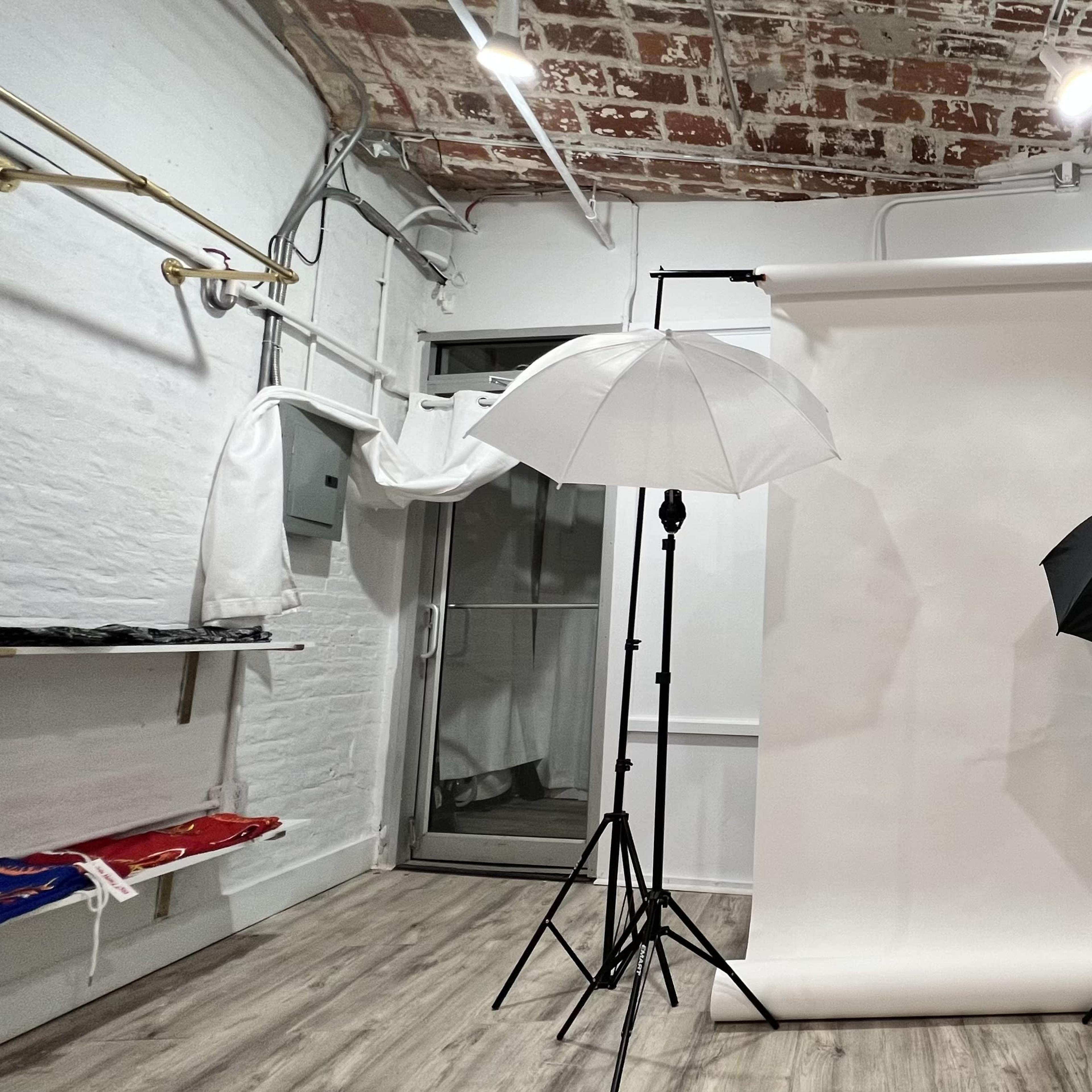 A photography studio with a white backdrop, two umbrellas (one white and one black) on tripods, and exposed brick walls.