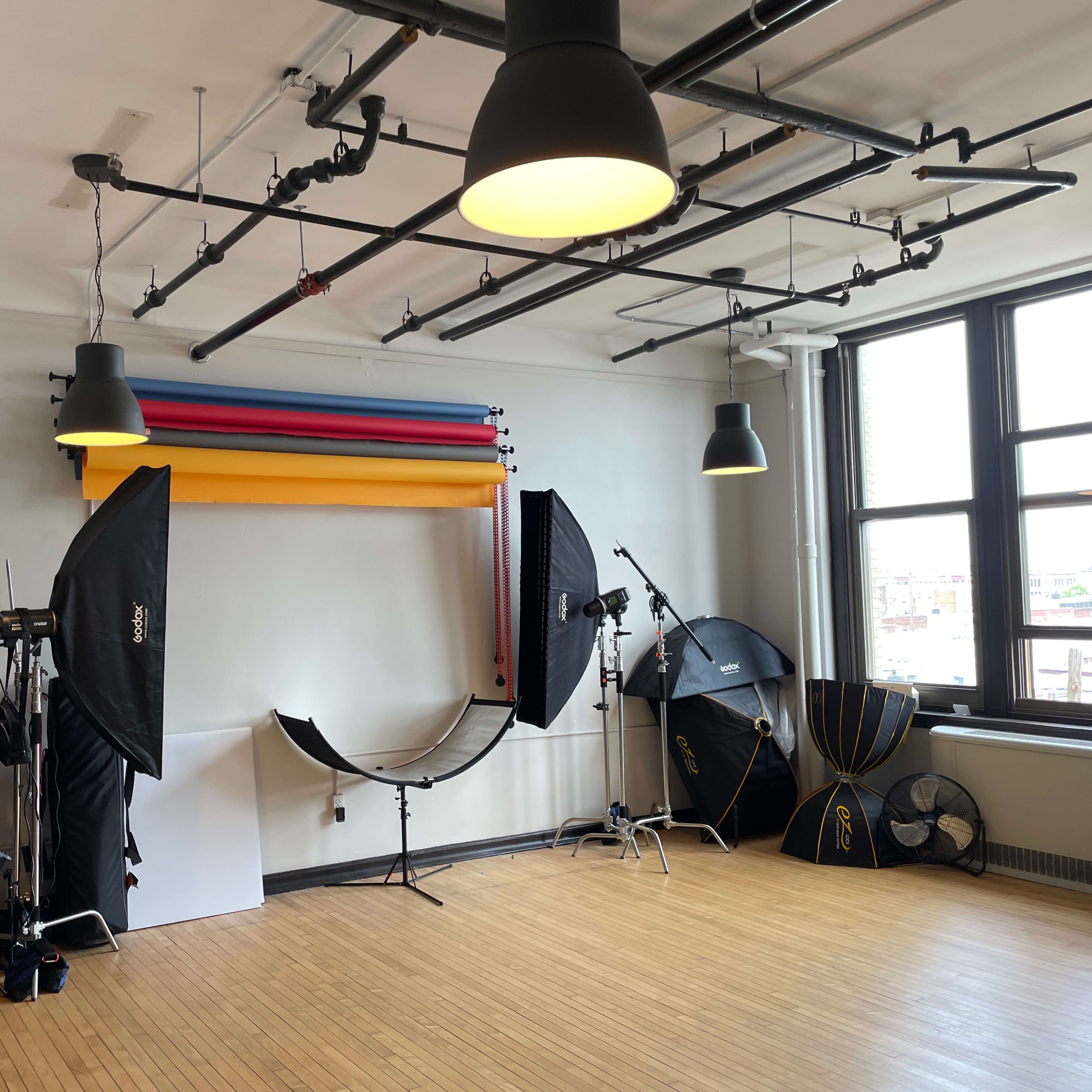 A spacious studio with a gray sofa, a brown chair, a black coffee table, and various photography equipment positioned around a large backdrop.