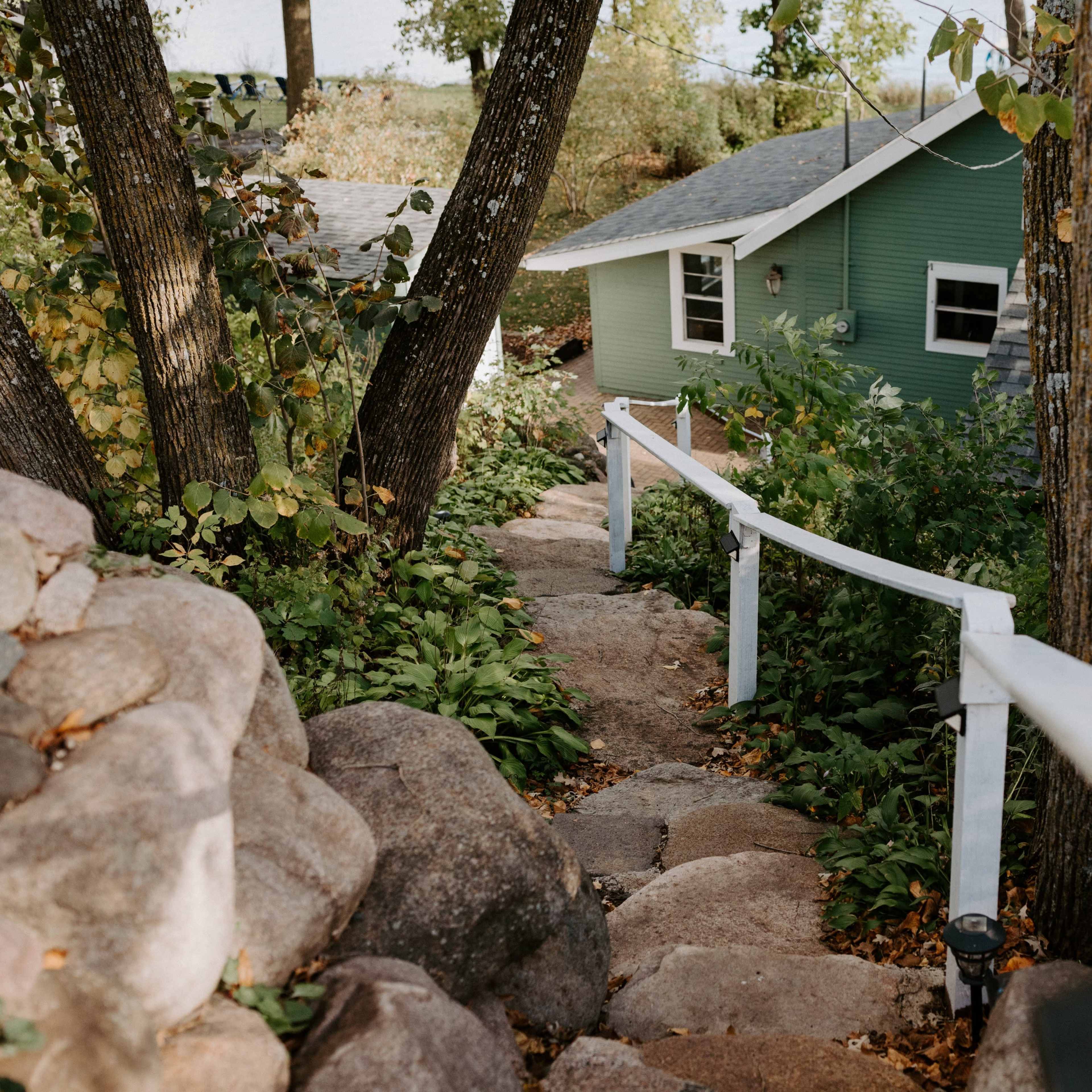 A stone pathway leads downwards through foliage to a green house surrounded by trees.