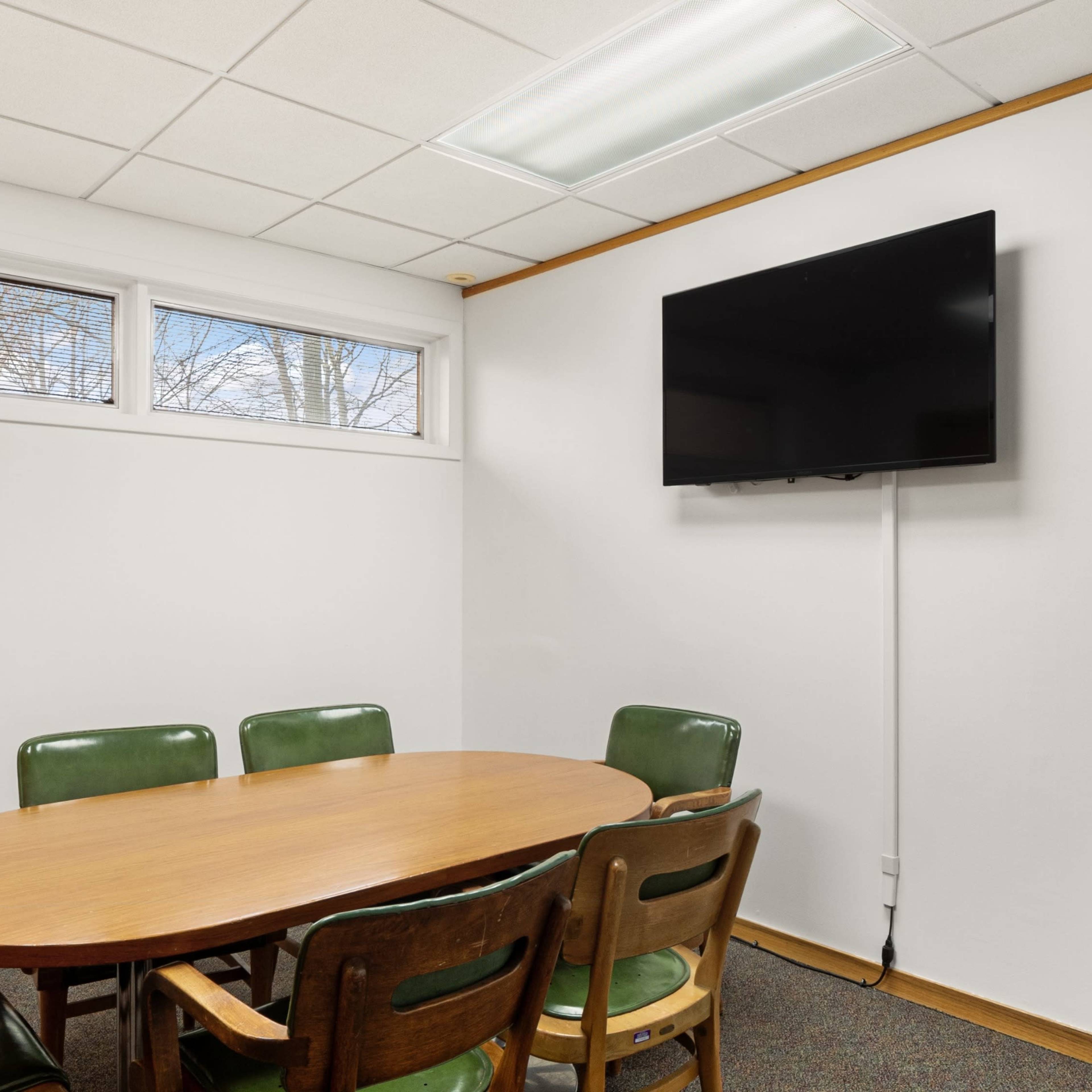 A conference room features a round wooden table surrounded by green and brown chairs, with a large television mounted on the wall and windows providing natural light.