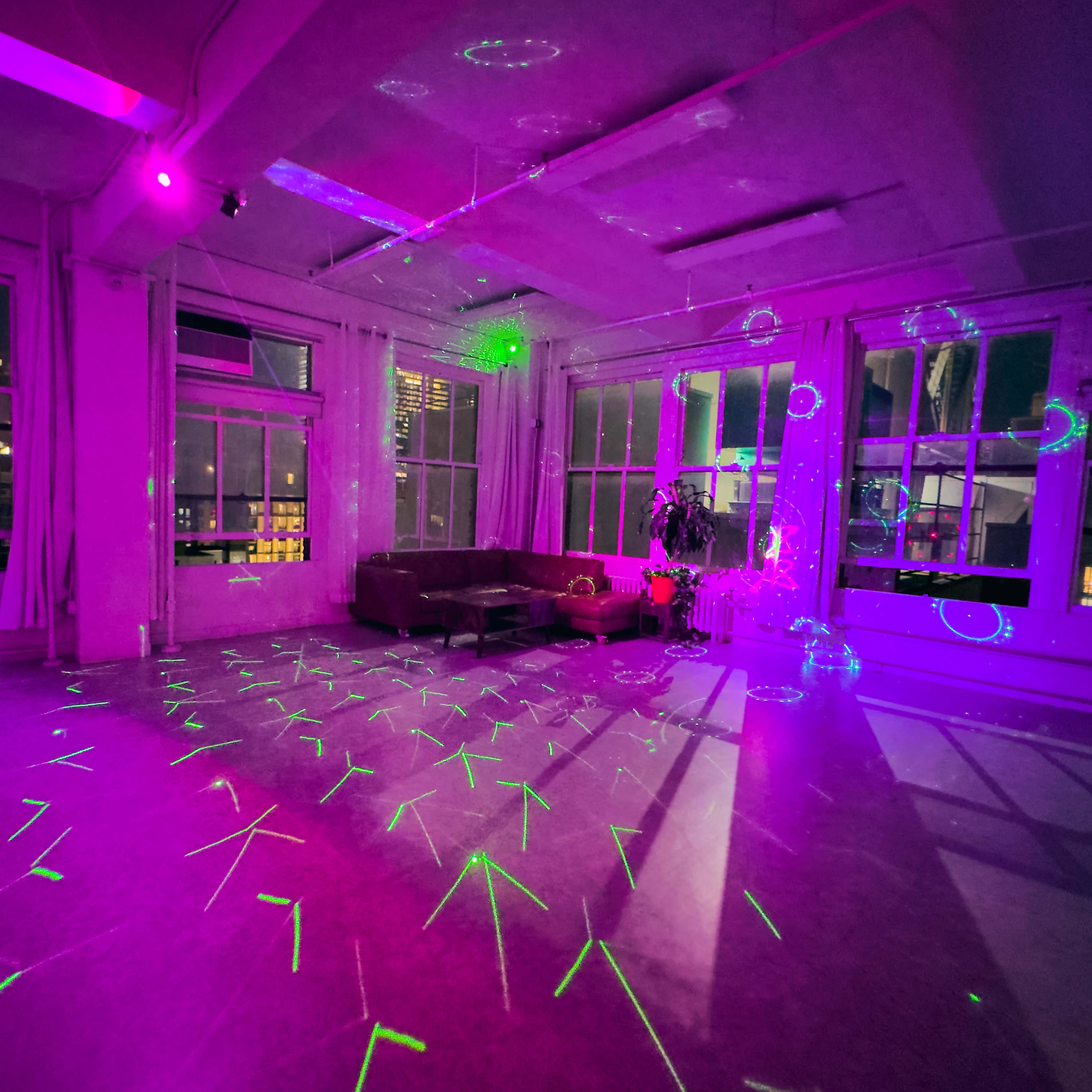 A brightly lit room features a couch and colorful laser patterns projected onto the floor, with large windows showing city lights outside.