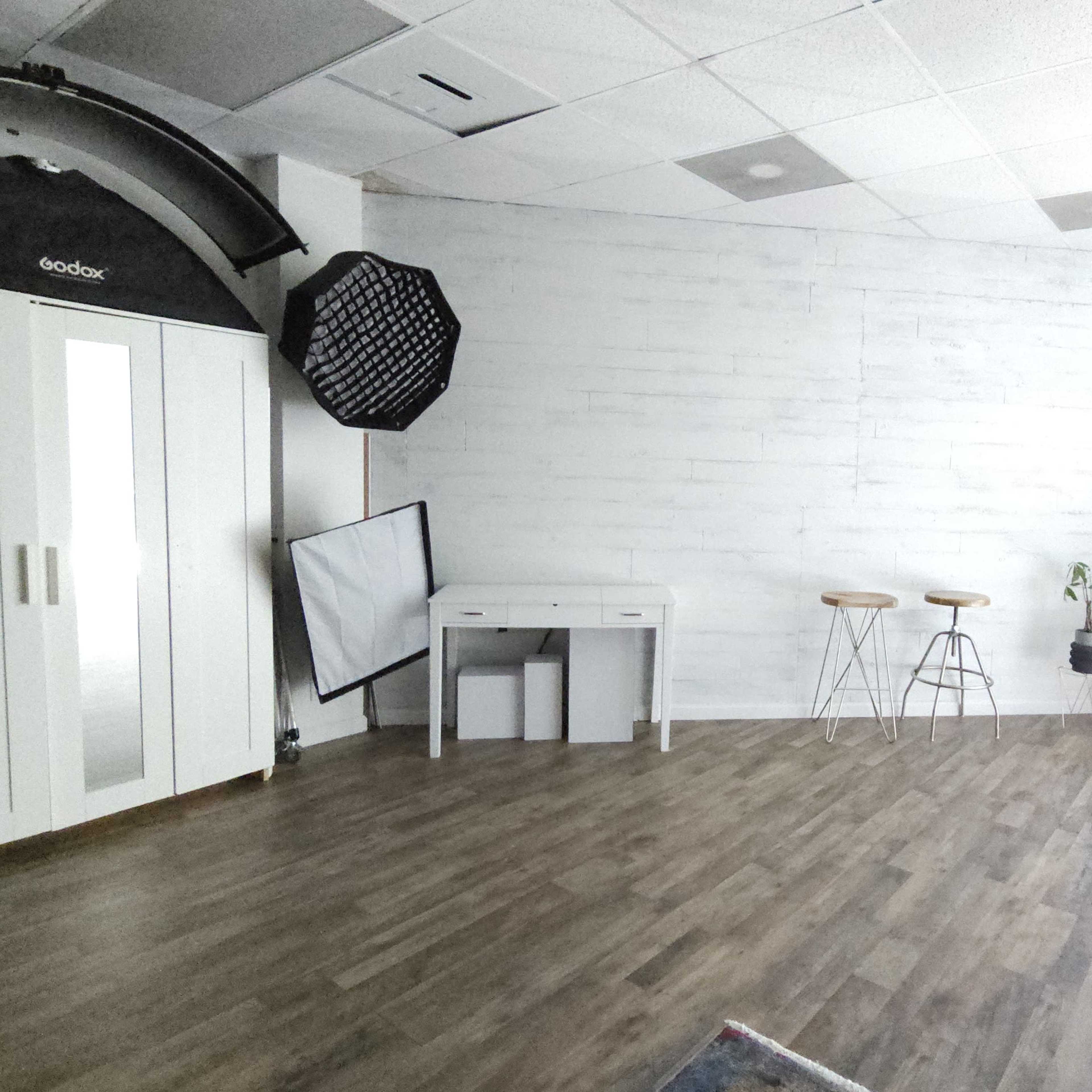 A spacious, minimalist studio with a light wooden floor, a white cabinet, a small desk, two stools, and large windows illuminating the area.