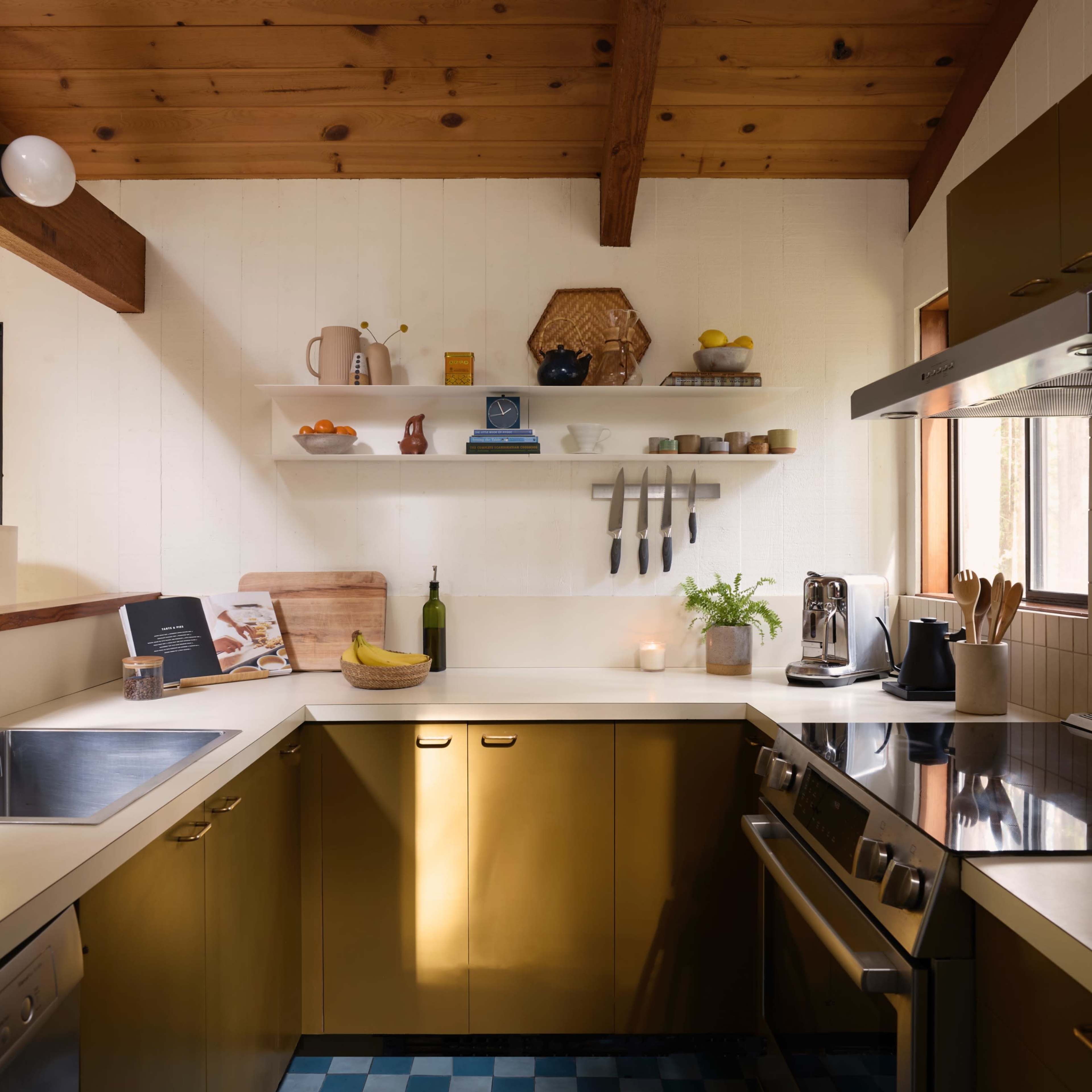 A modern kitchen features green cabinetry, a stainless steel stove, a large sink, and wooden beams above a countertop with various kitchen items and a window letting in natural light.