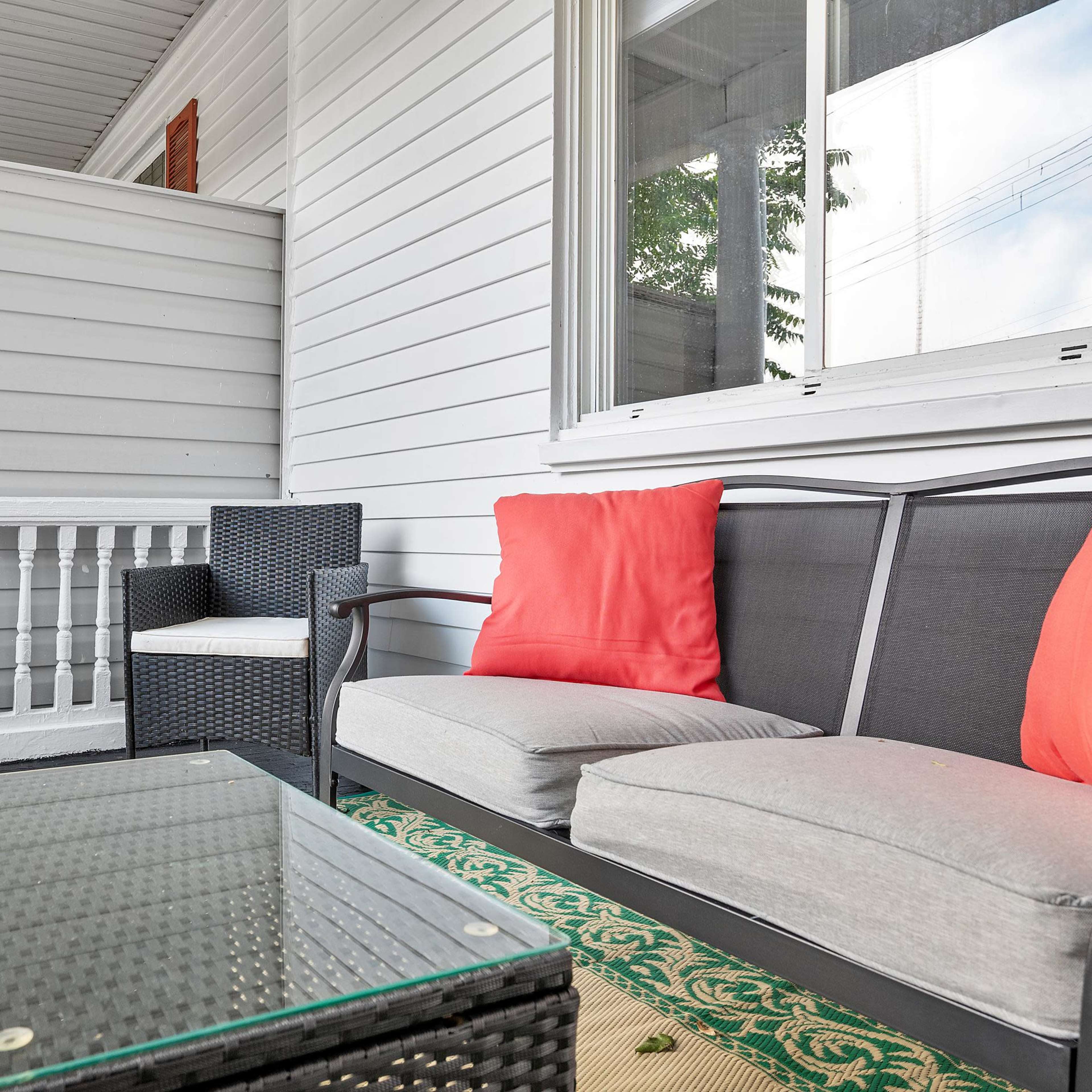 A small outdoor seating area with black wicker furniture, including two chairs and a couch, arranged around a glass-topped table on a patterned rug.