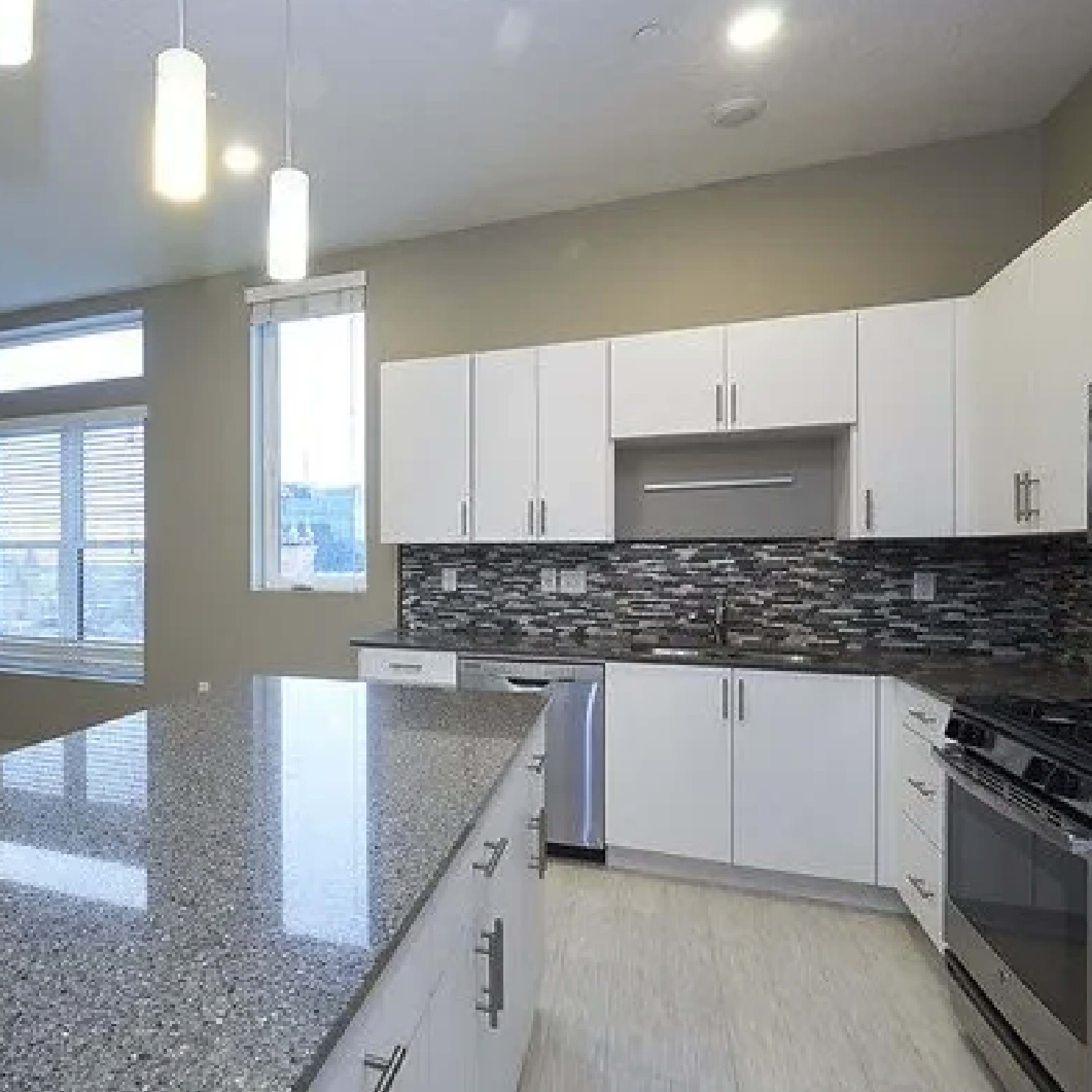 A modern kitchen with white cabinetry, a large granite countertop, and a tiled backsplash, complemented by pendant lighting.