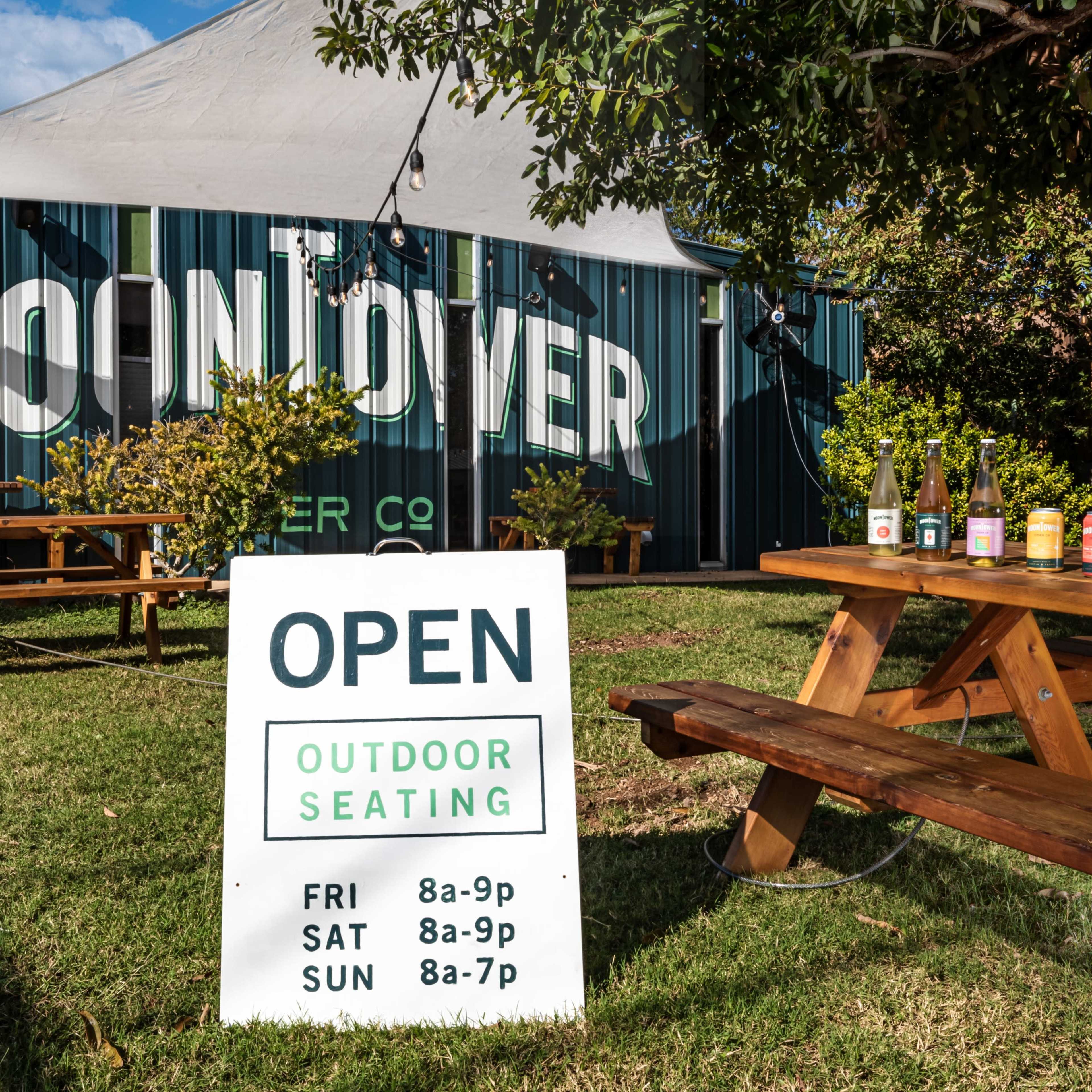 A sign indicating "Open" for outdoor seating, with a picnic table displaying drinks in front of a building labeled "Moon Over" in a green and white color scheme.