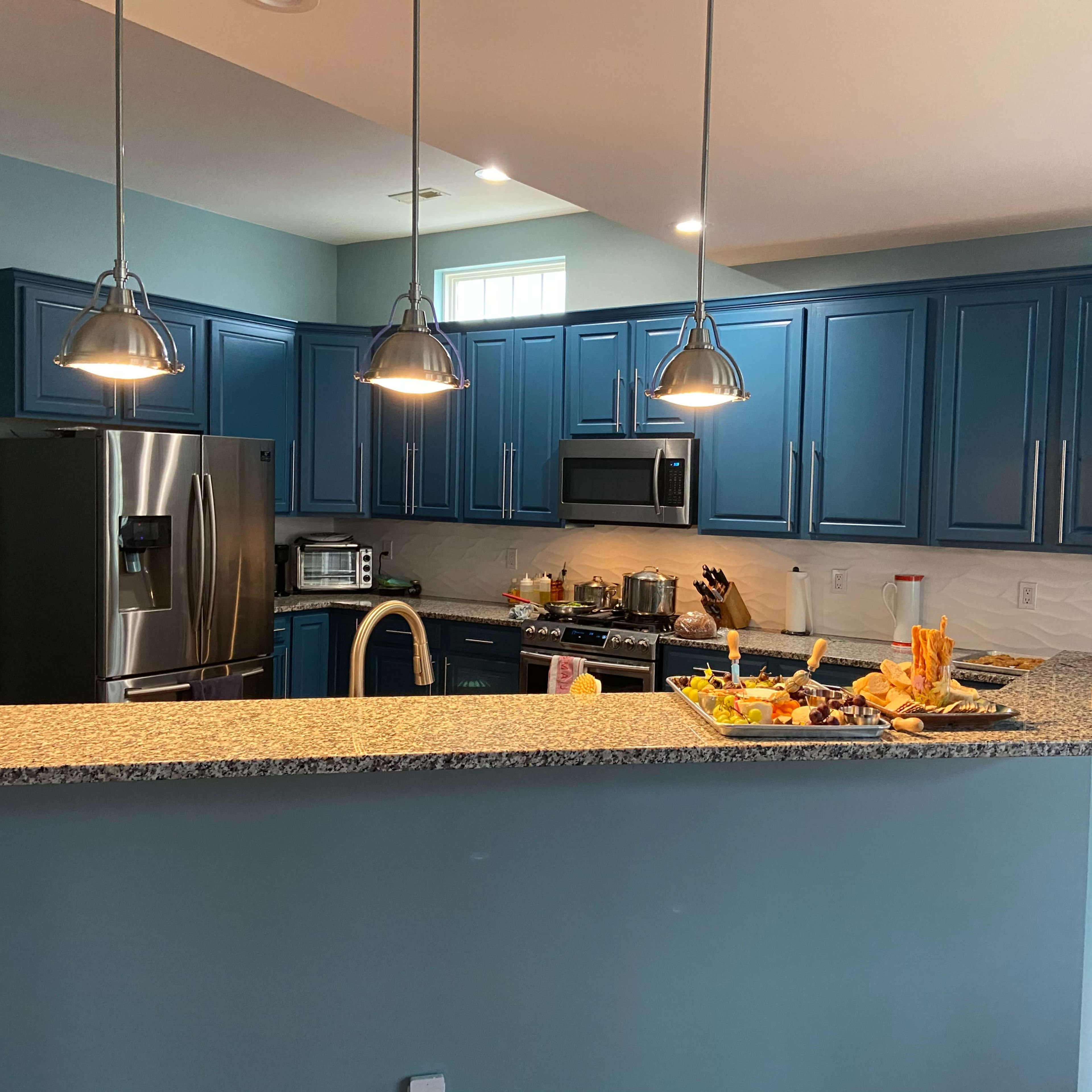 A modern kitchen with blue cabinetry, granite countertops, and pendant lighting, featuring a variety of food items on the island.
