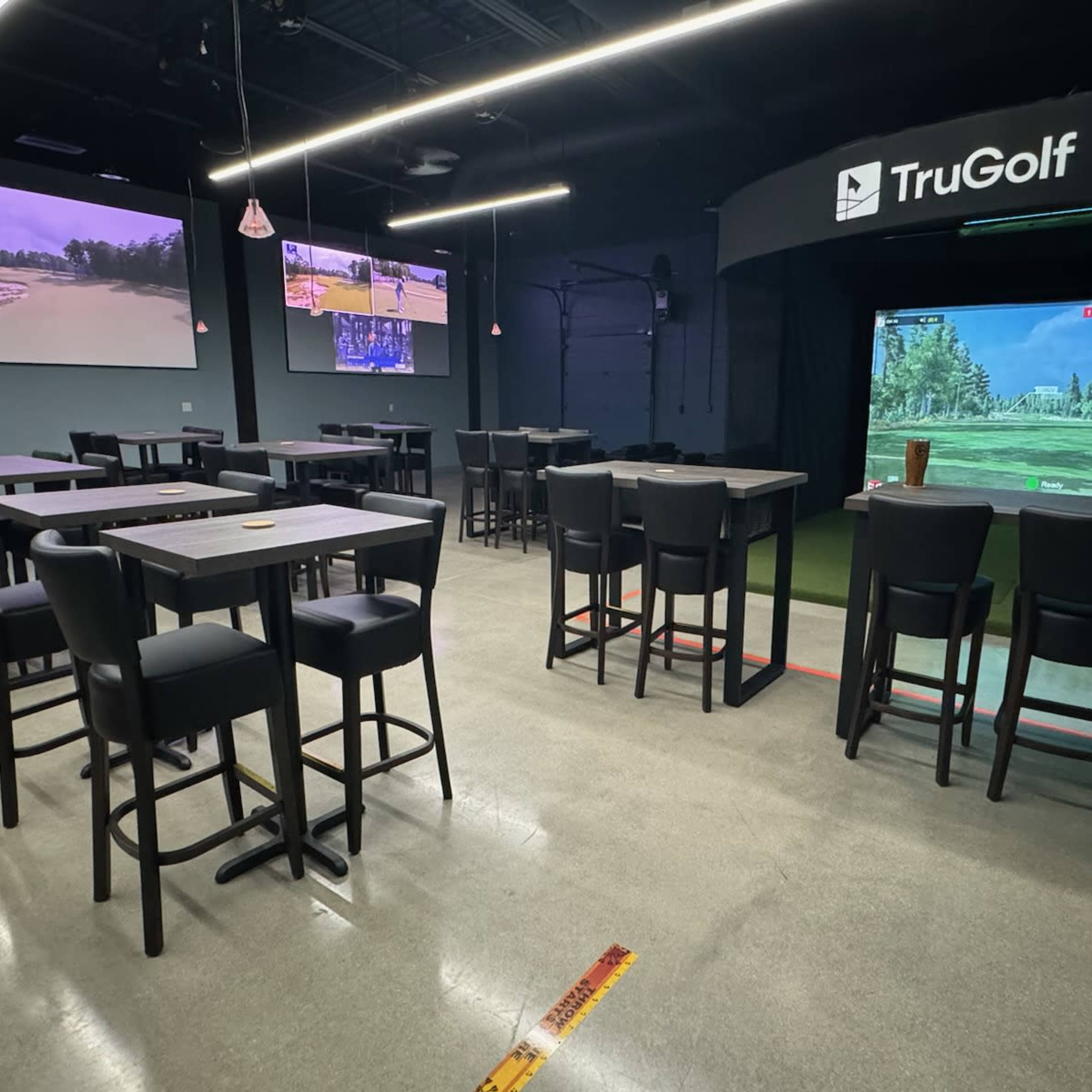 An indoor golf simulation facility with multiple screens displaying golf courses, surrounded by high tables and bar stools.