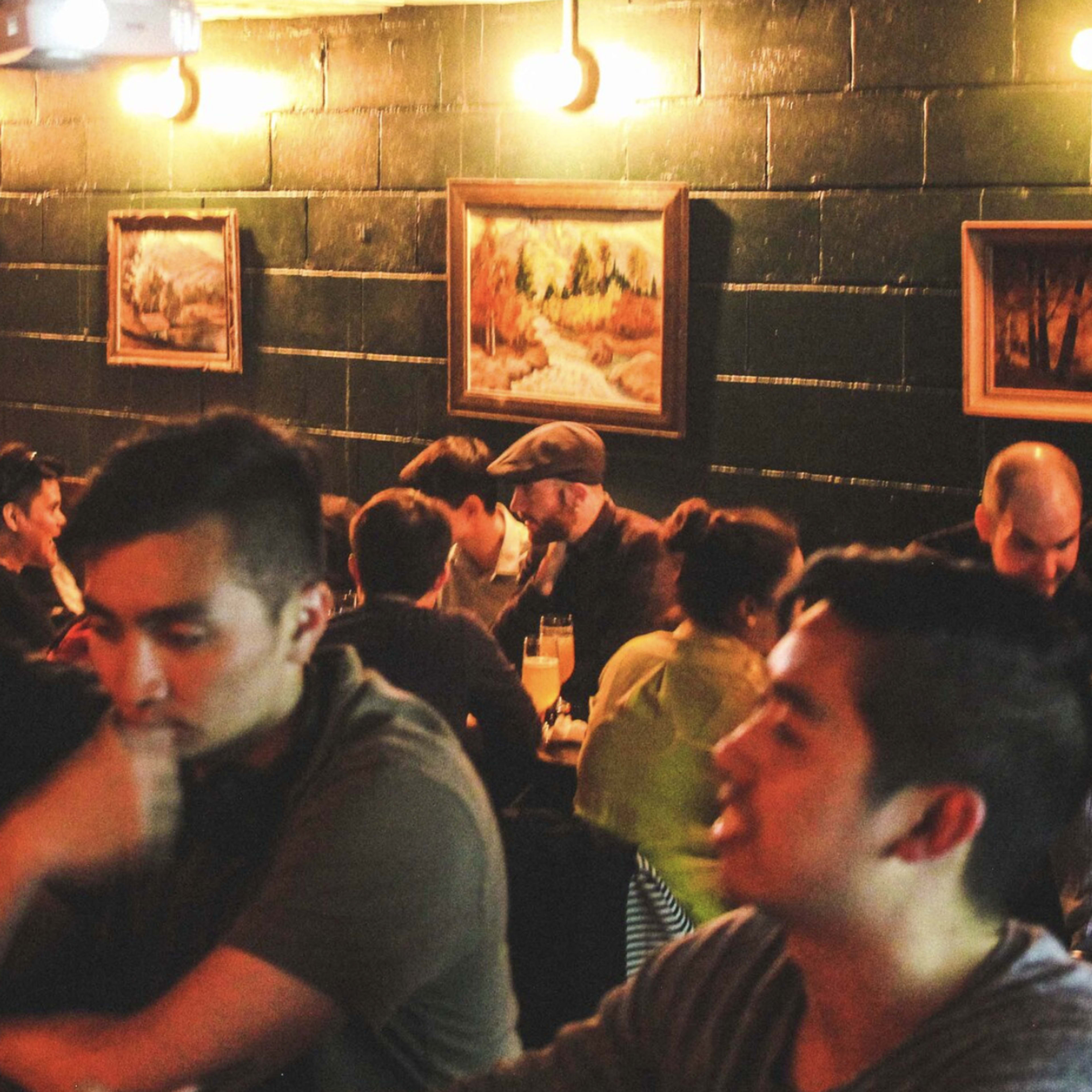 A busy bar interior features patrons seated at a long wooden table, engaged in conversation and surrounded by framed landscape paintings on the walls.