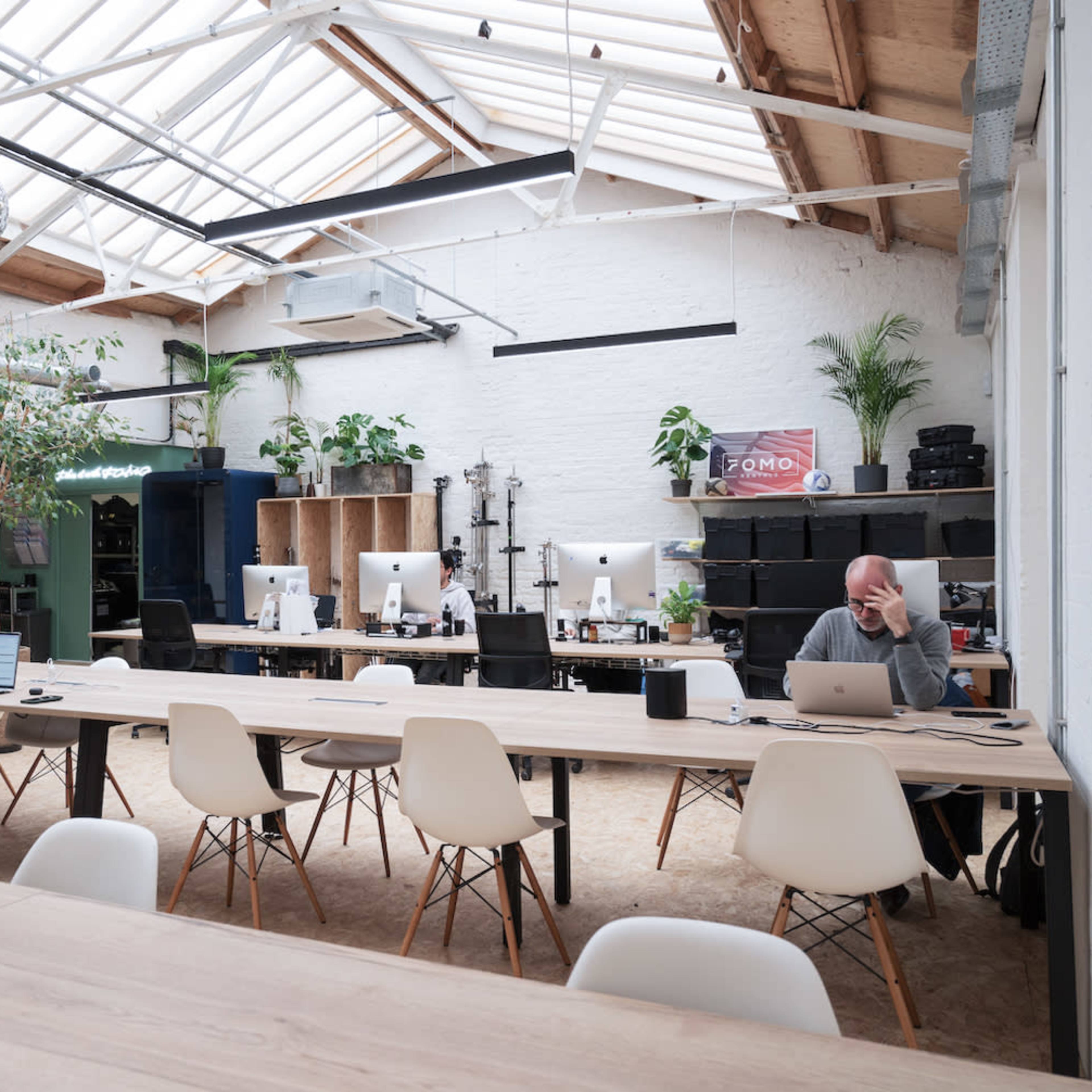 A spacious coworking space features several desks with computers, a large communal table, and plants, all under a glass ceiling.