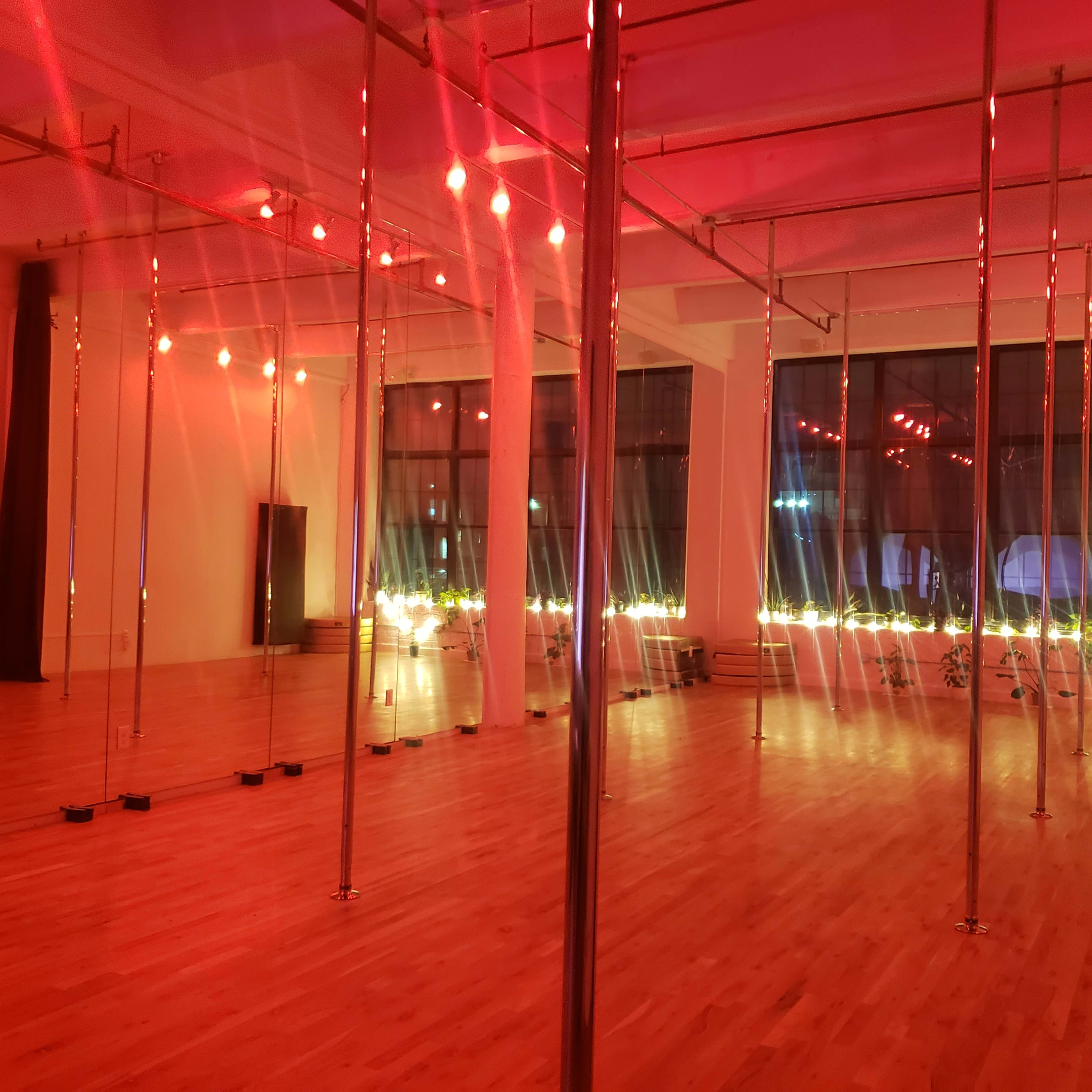 A dance studio with multiple polished poles and red lighting, featuring wooden flooring and large windows illuminated by soft lights.