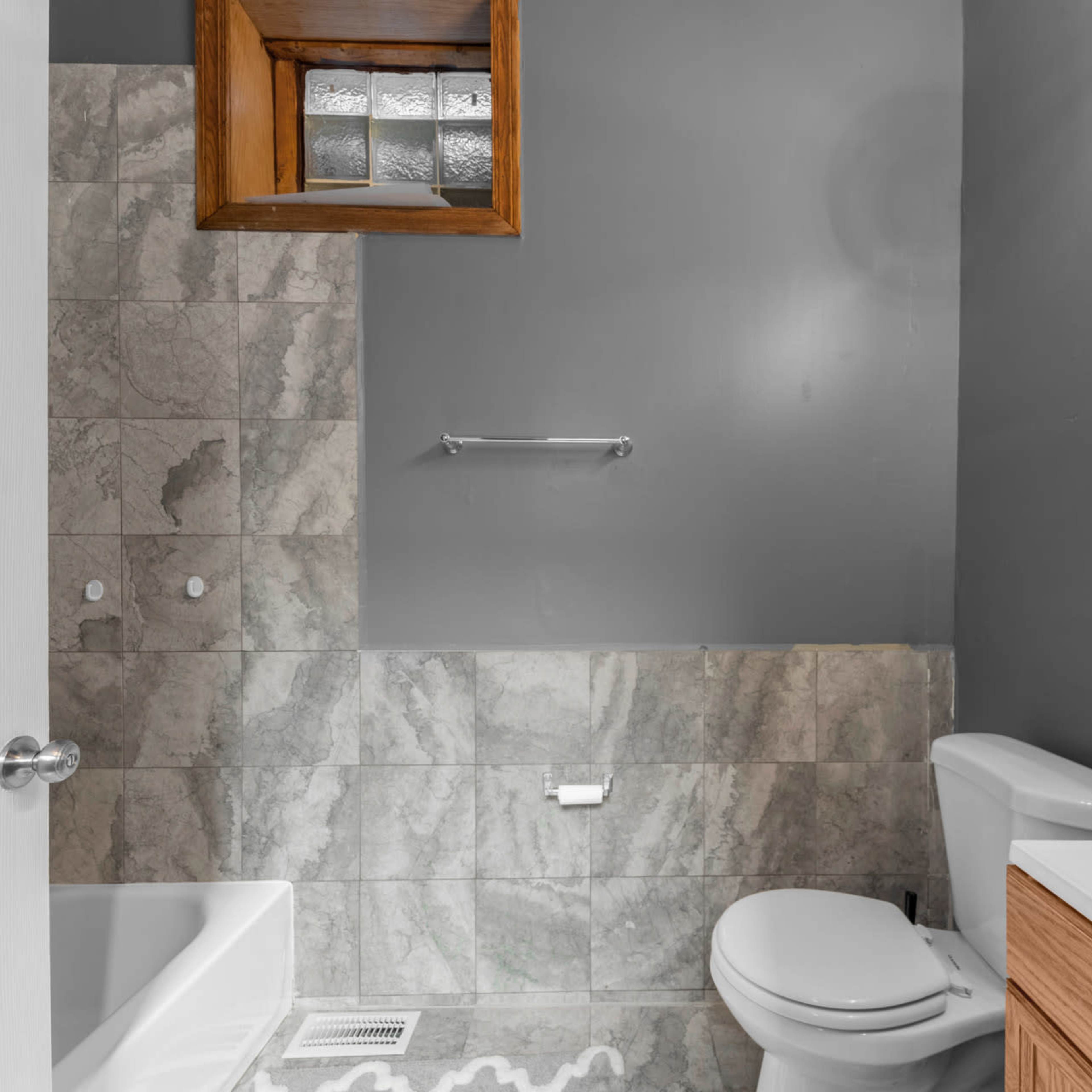 A bathroom with gray walls, a marble-patterned tile floor, a white bathtub, a toilet, and a wooden vanity with a mirror.