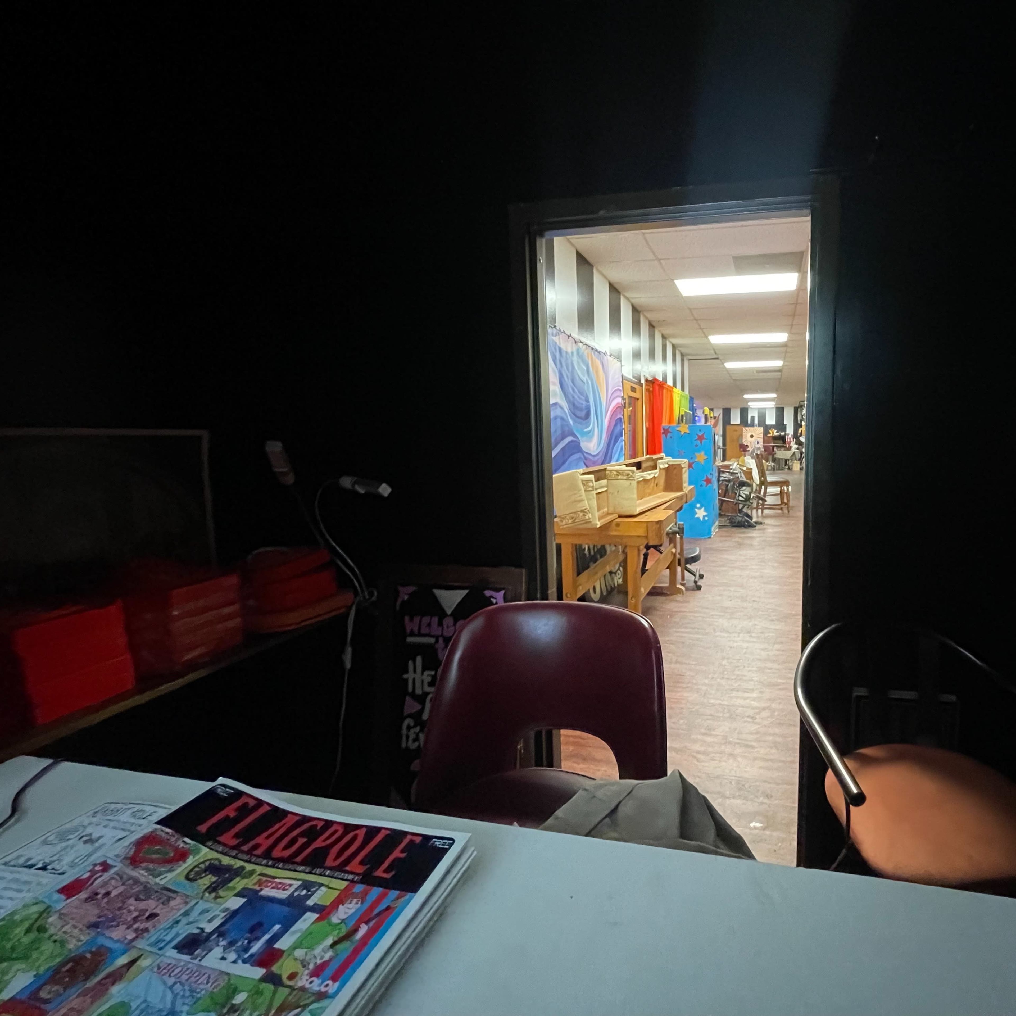 A dark room with a table and two chairs leads to a brightly lit hallway filled with colorful artwork and tables in the background.