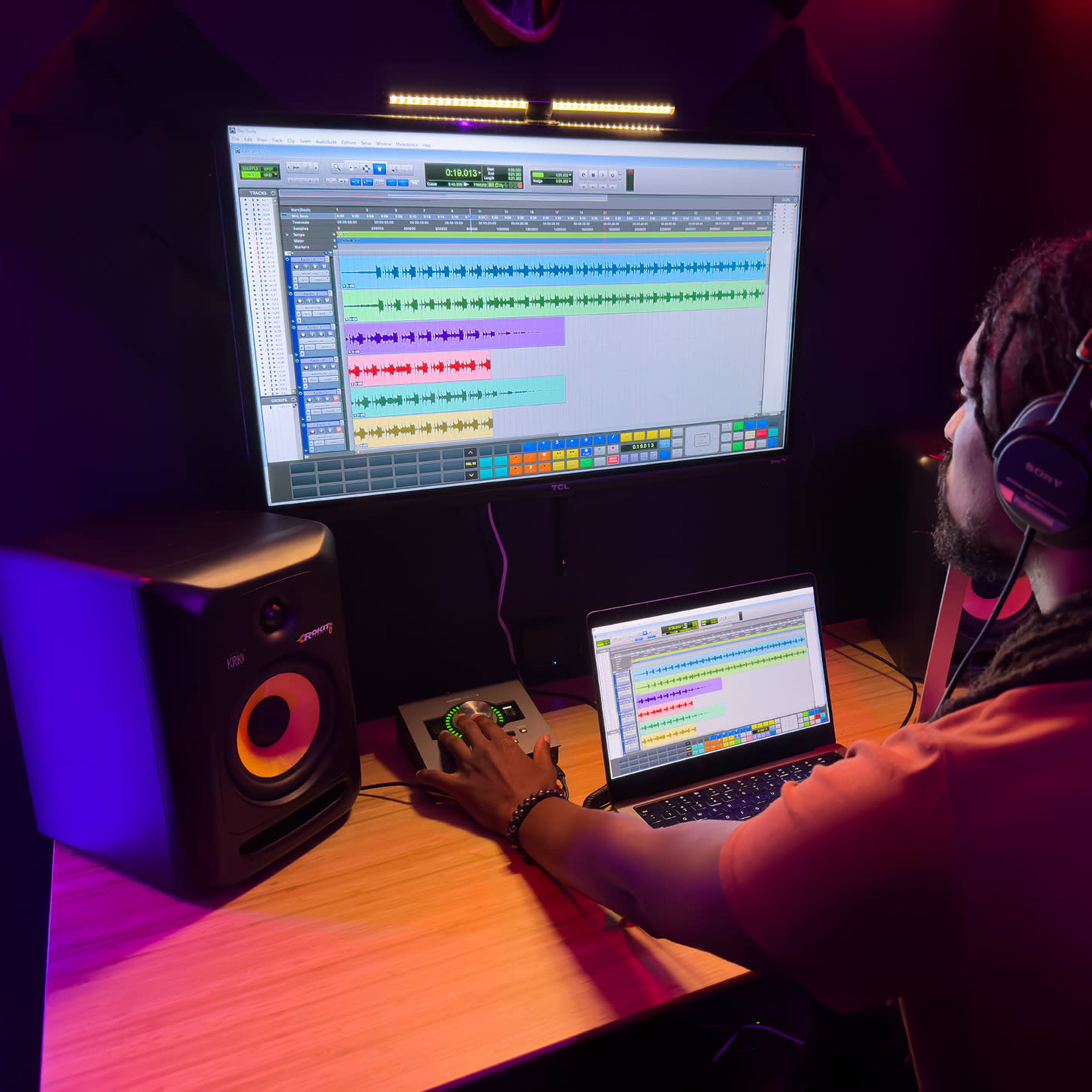 A person is working at a desk with a laptop and a large monitor displaying music production software, while speakers are positioned on either side.