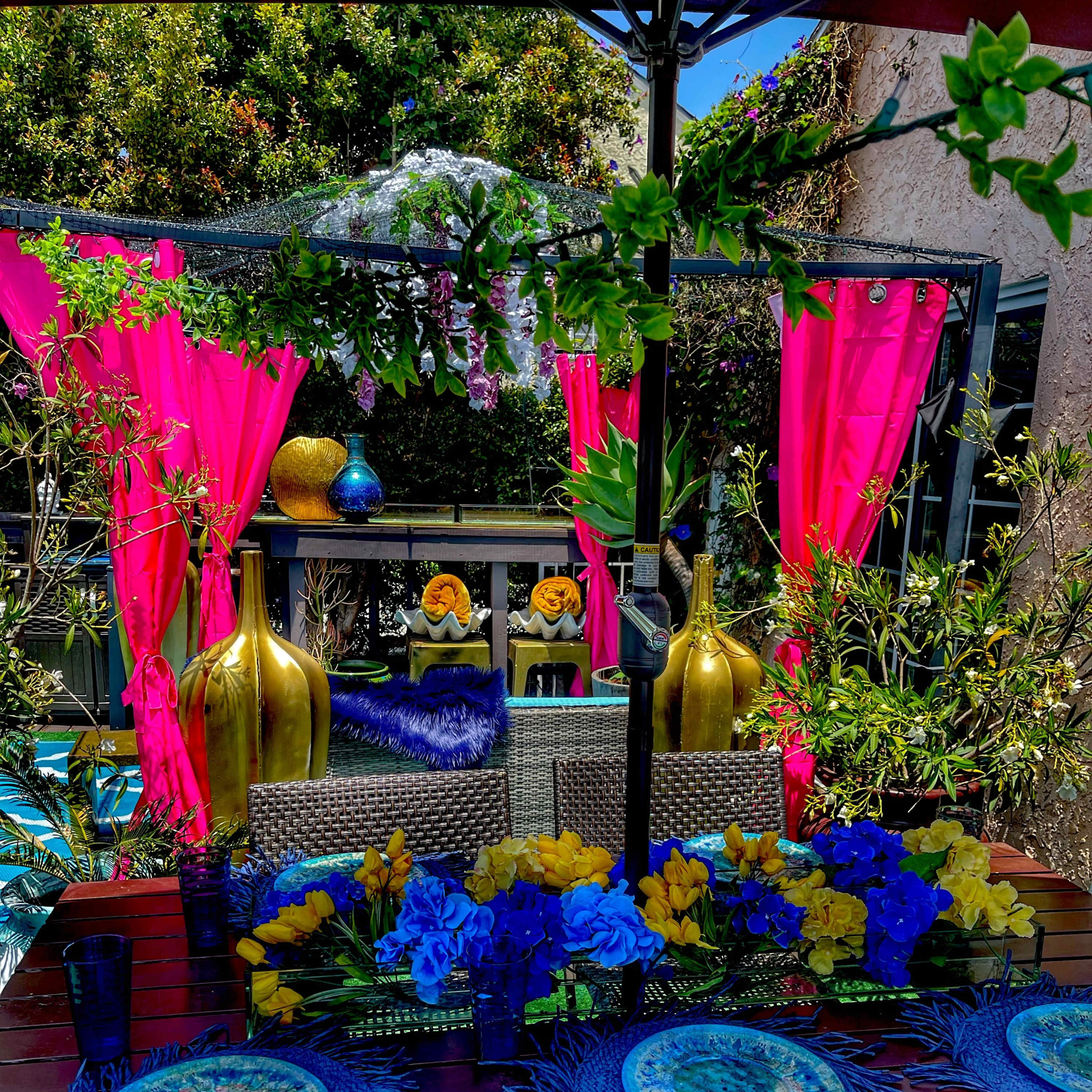 A vibrant outdoor dining area features a table set with blue and yellow floral arrangements, colorful place settings, and pink drapes hanging in the background.