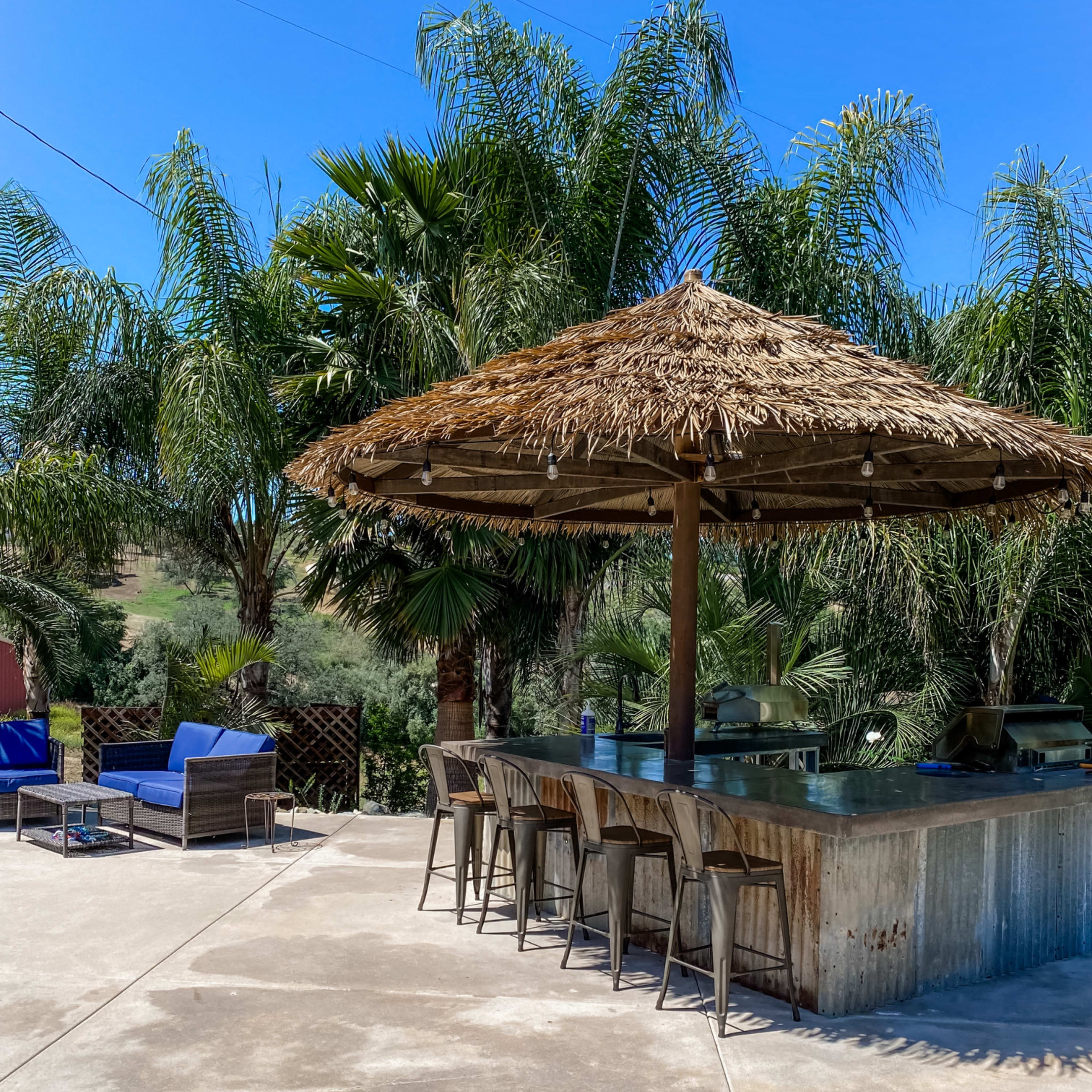 A shaded outdoor bar with high stools and a thatched roof is surrounded by palm trees and lounge chairs on a concrete patio.