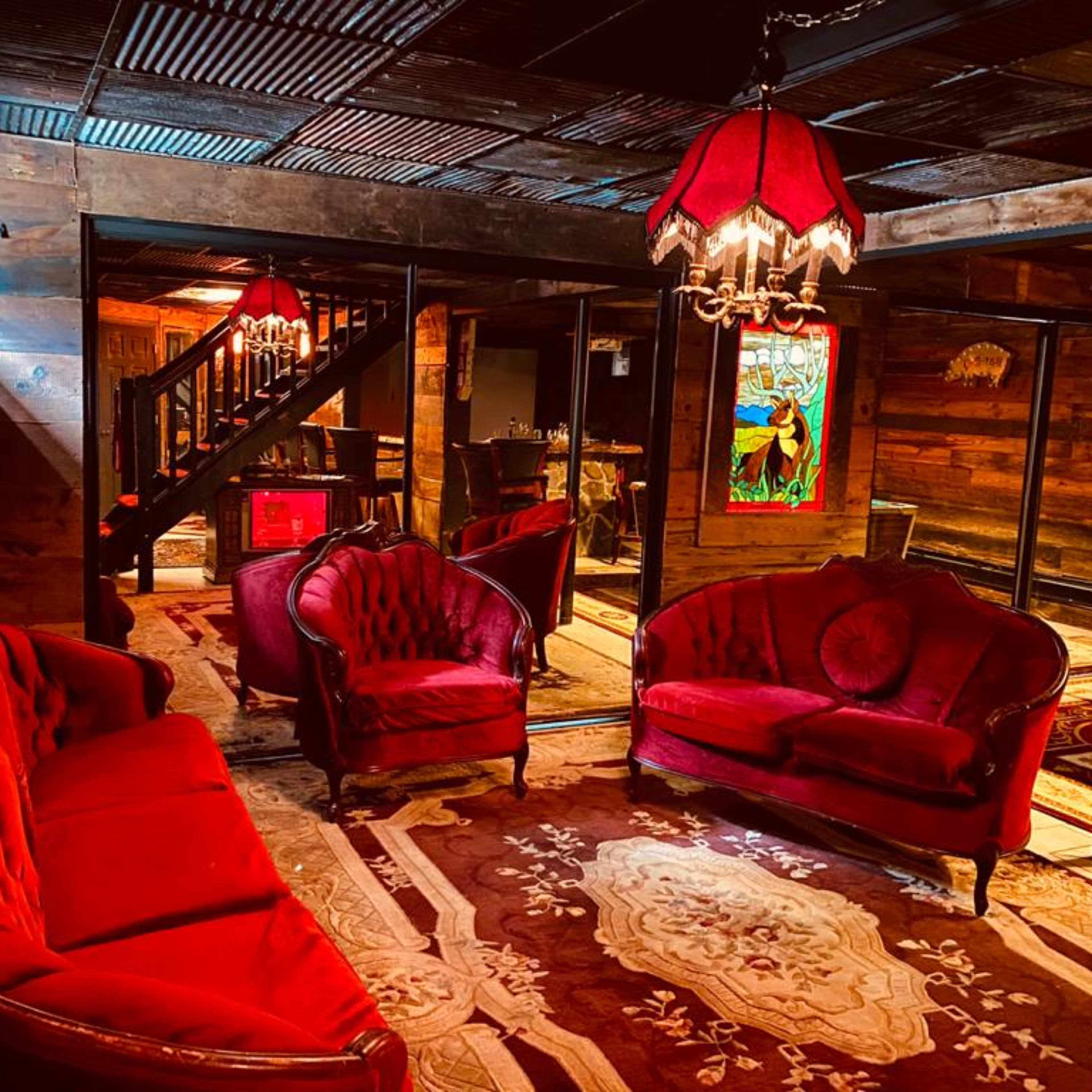 A rustic lounge area featuring red velvet furniture, patterned rugs, and a stained glass window, all illuminated by vintage lamps.