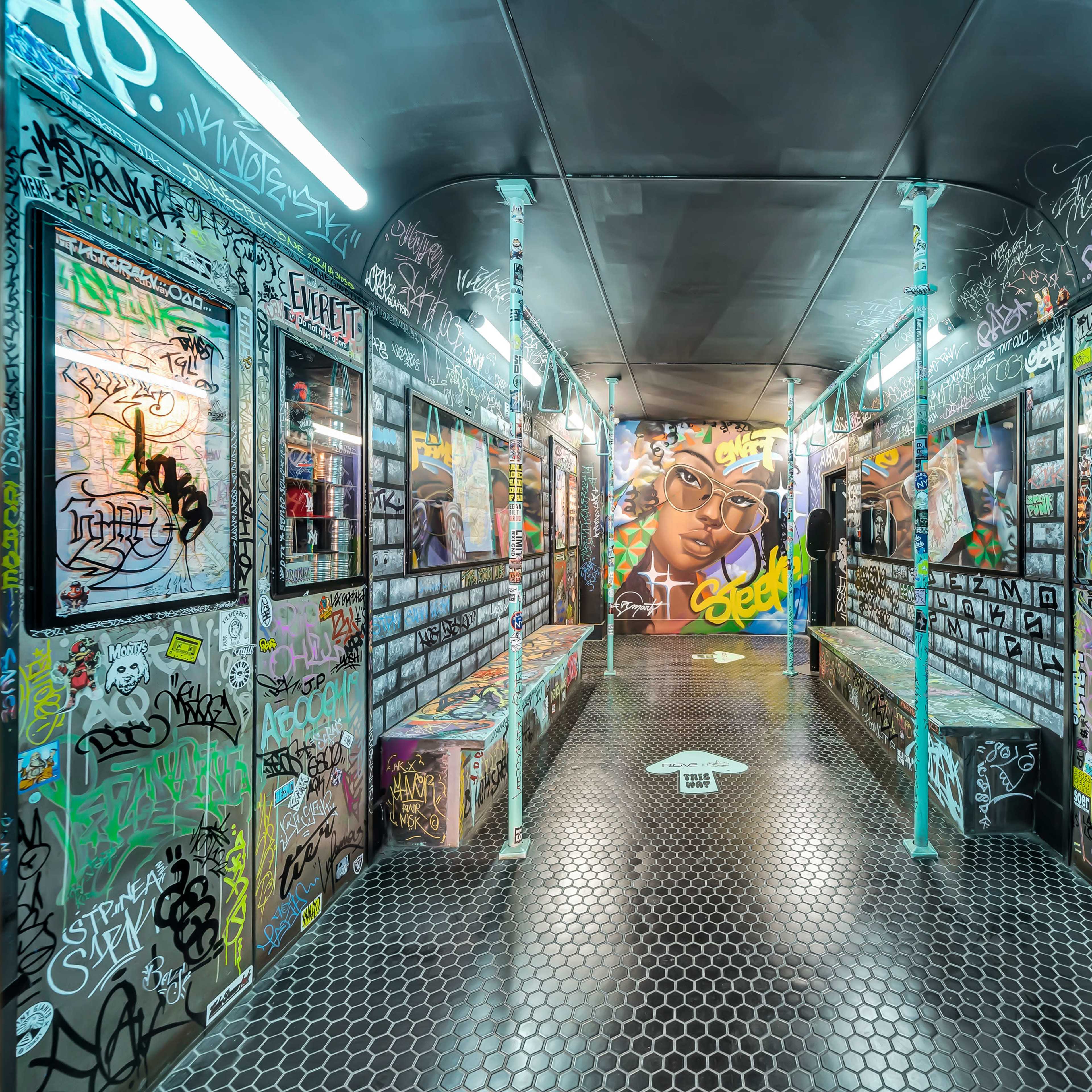 A narrow, graffiti-covered hallway with mirrors on the walls and benches on either side, featuring a large mural of a face at the end.