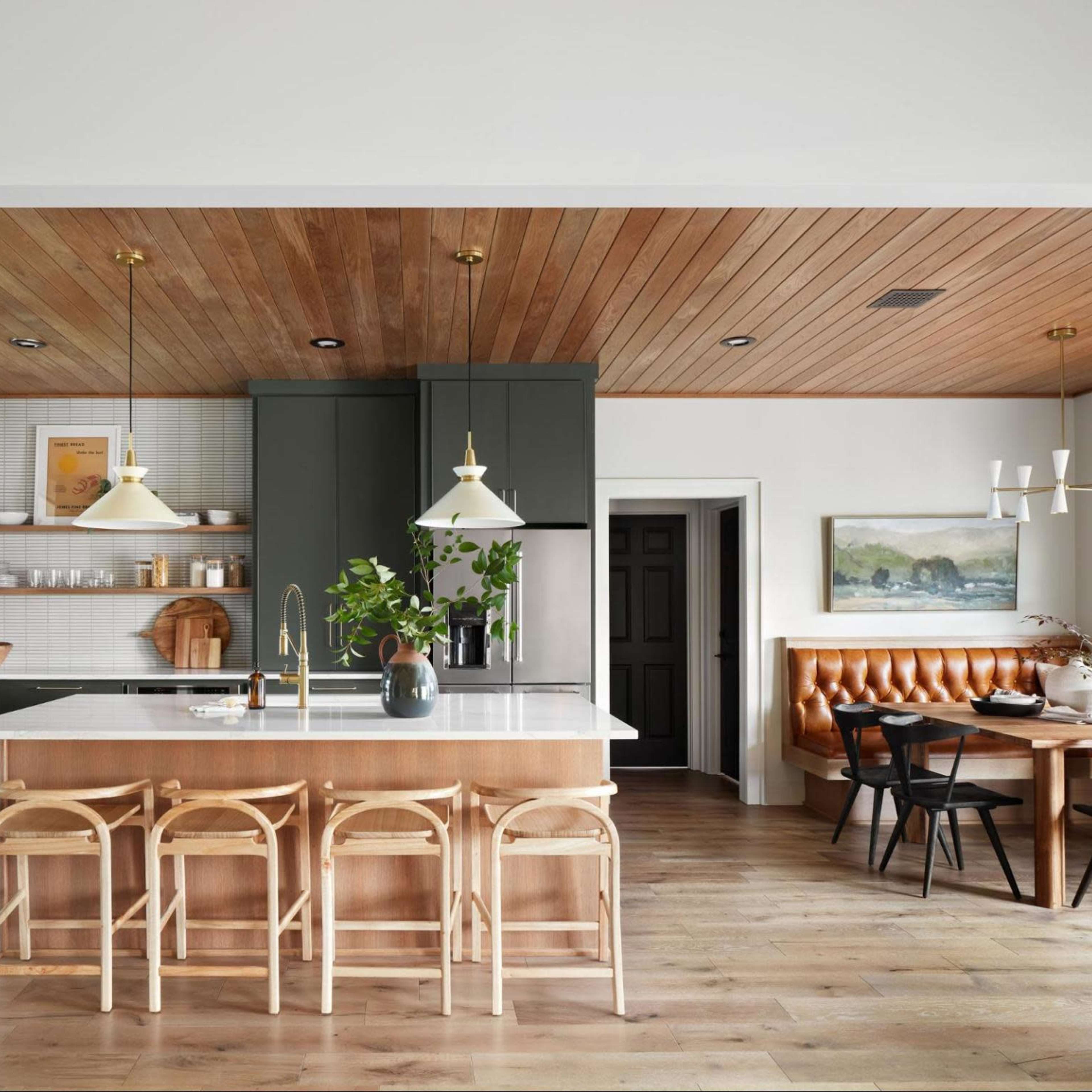 A modern kitchen features a central island with bar stools, green cabinetry, wooden ceilings, and a dining area with a round table and leather seating.