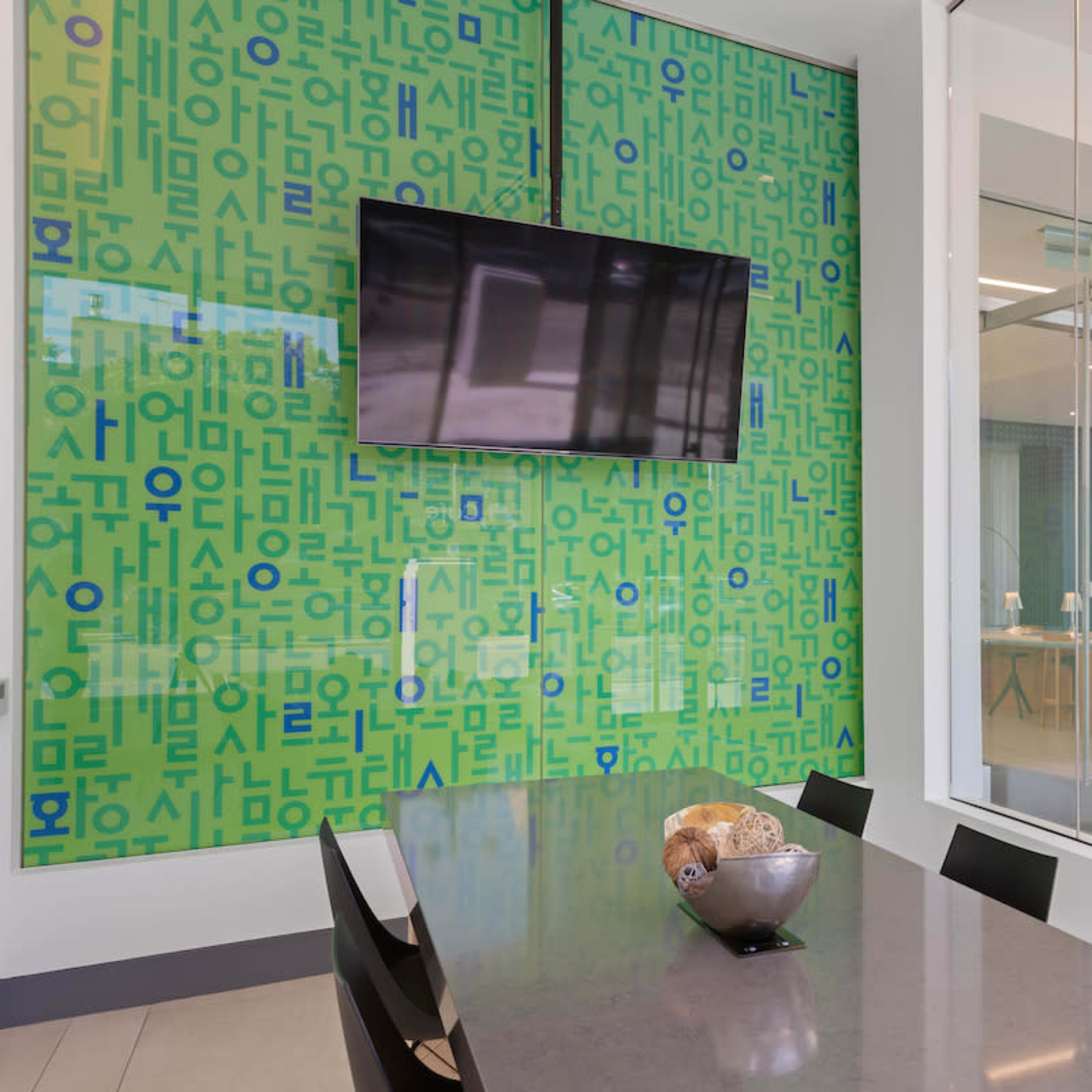 A modern conference room with a large table, a television mounted on a green wall covered in text, and glass doors leading to another room.