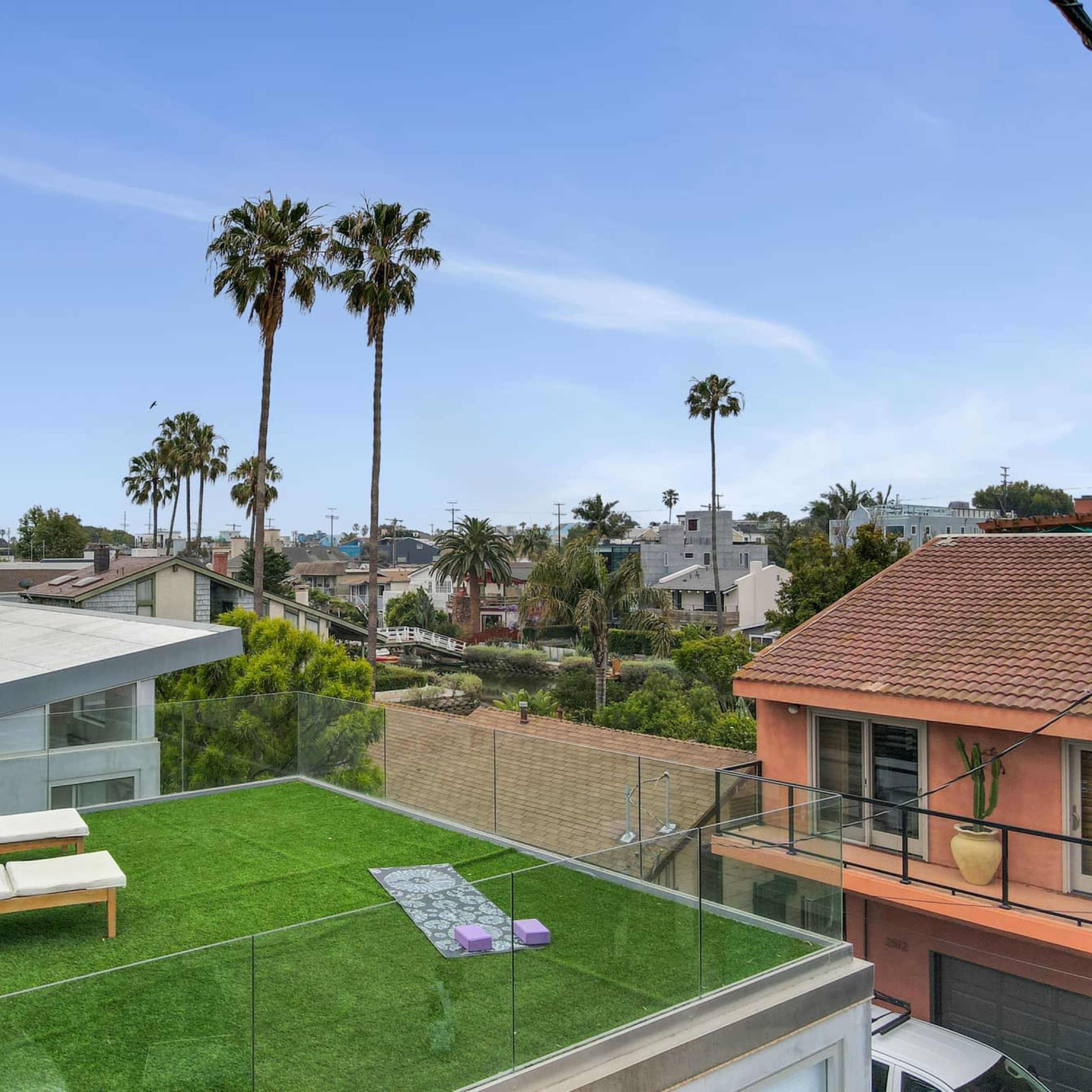 A rooftop deck with artificial grass, two lounge chairs, and palm trees in the background overlooking residential buildings.
