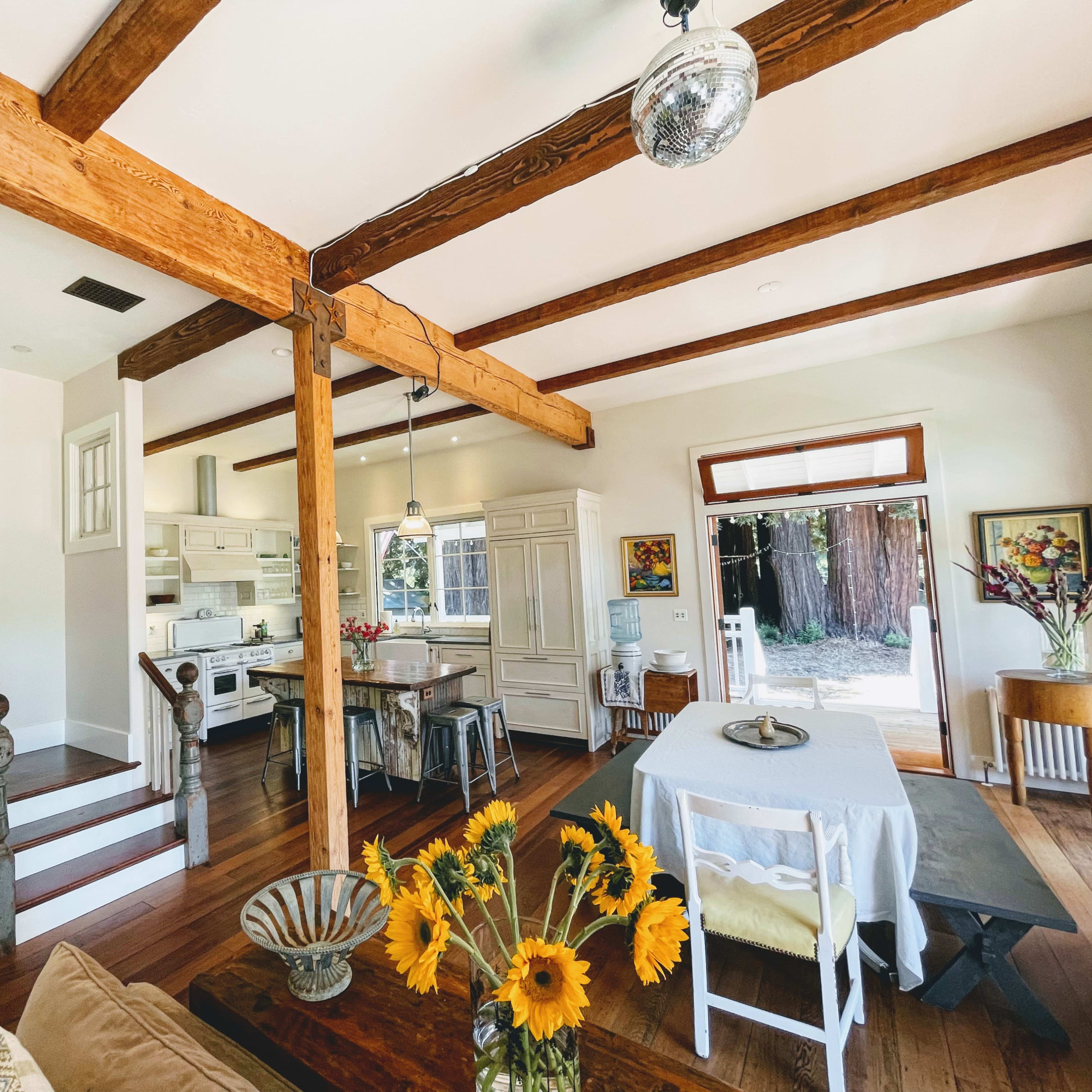 A spacious living area features wooden beams, a dining table with sunflowers, and a staircase leading to an upper level.
