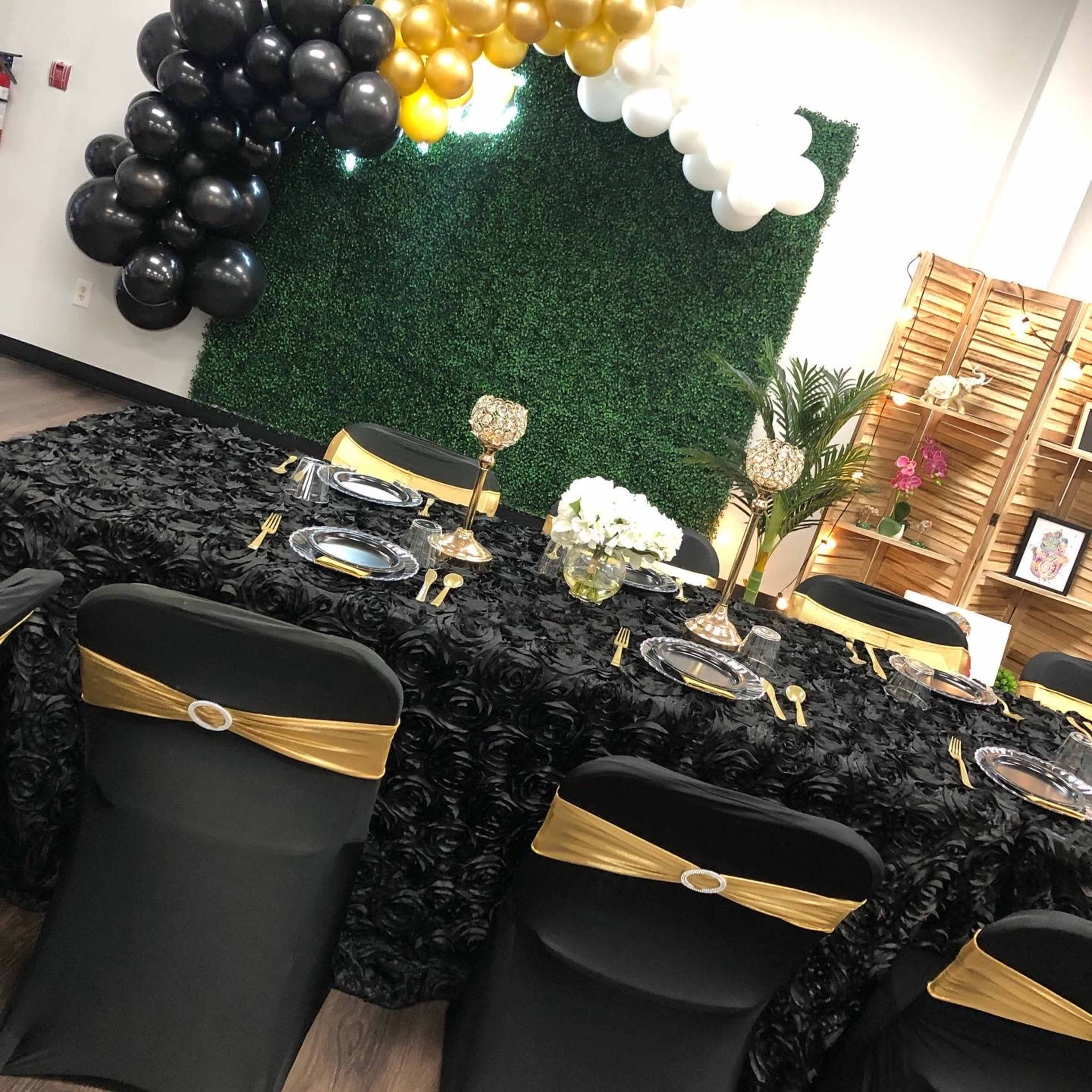 A decorated dining table is set with black and gold accents, flower centerpieces, and a backdrop of green foliage and balloons.