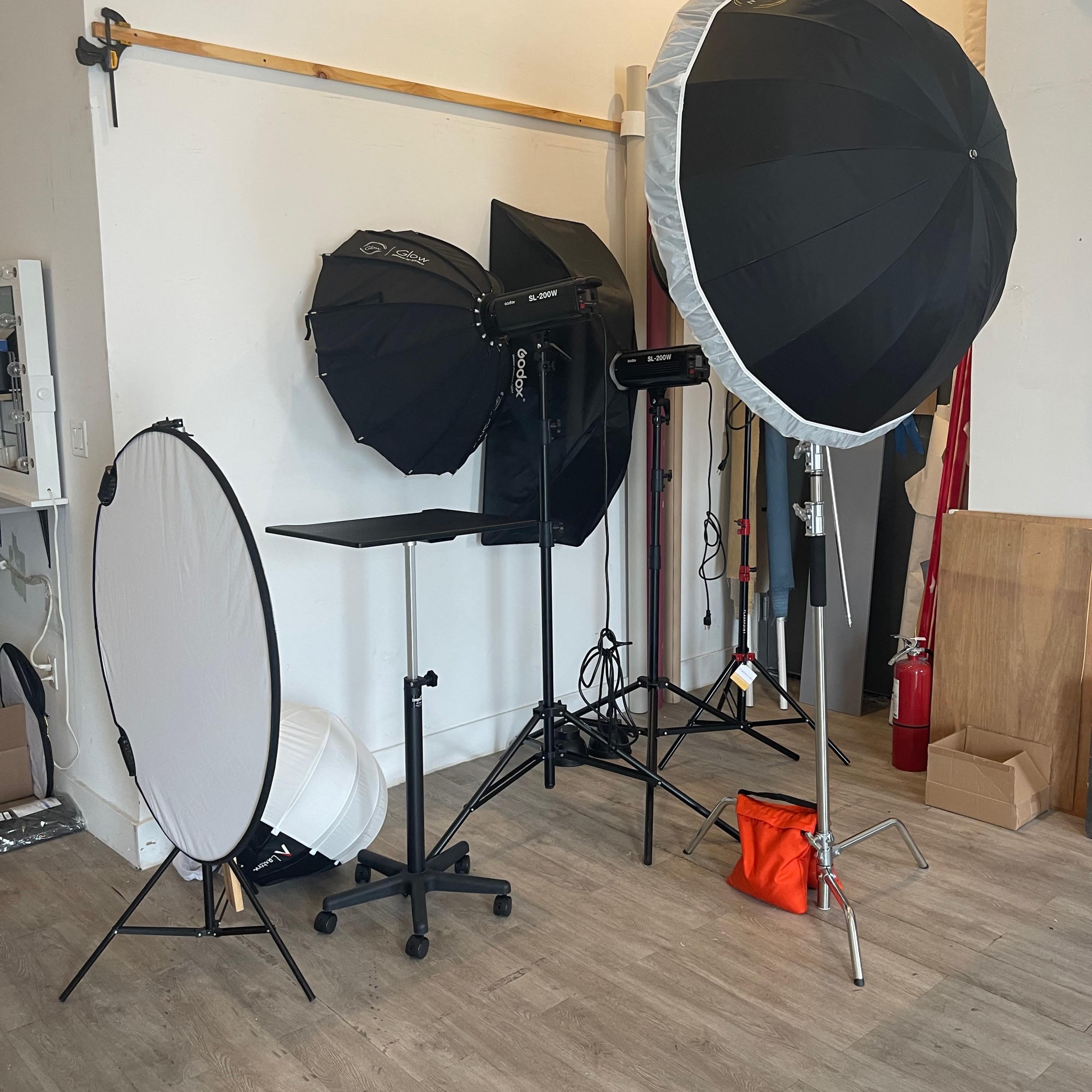 A photography studio setup with various lighting equipment, including softboxes, reflectors, and light stands, arranged against a wall.