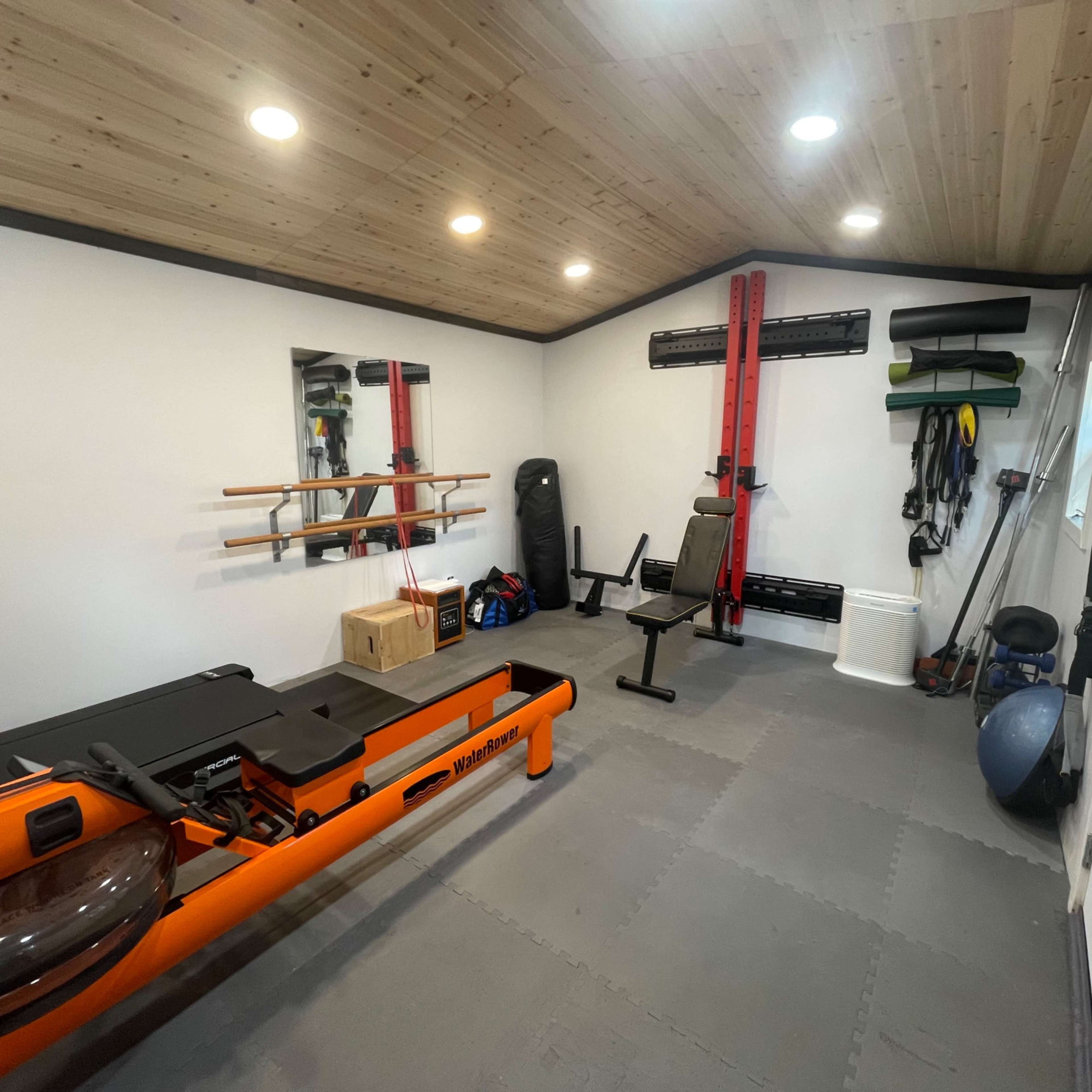 A home gym with fitness equipment, including a rowing machine, weights, and exercise mats, in a well-lit room with wooden ceiling panels.