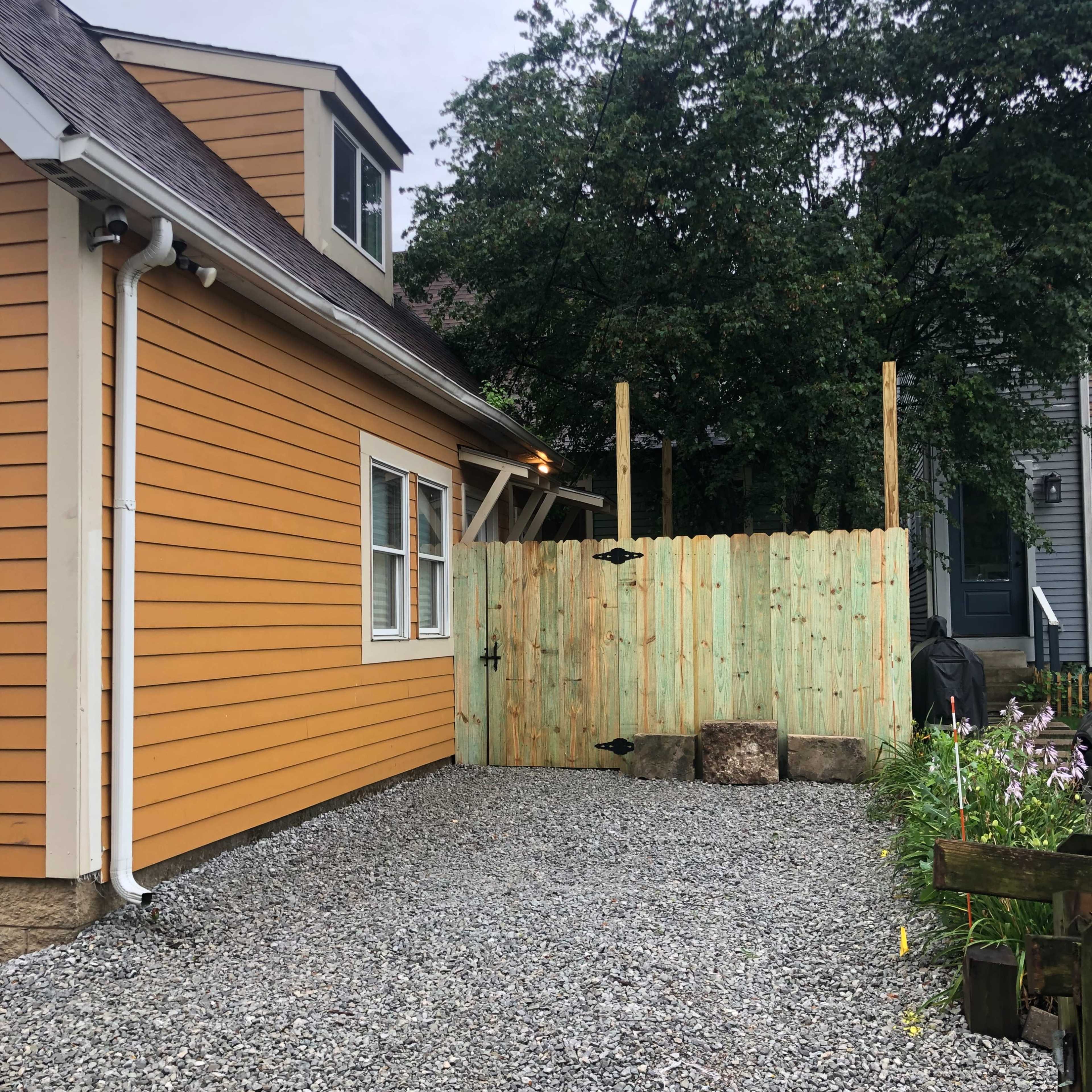 A gravel driveway leads to a house with orange siding, adjacent to a wooden fence and greenery.