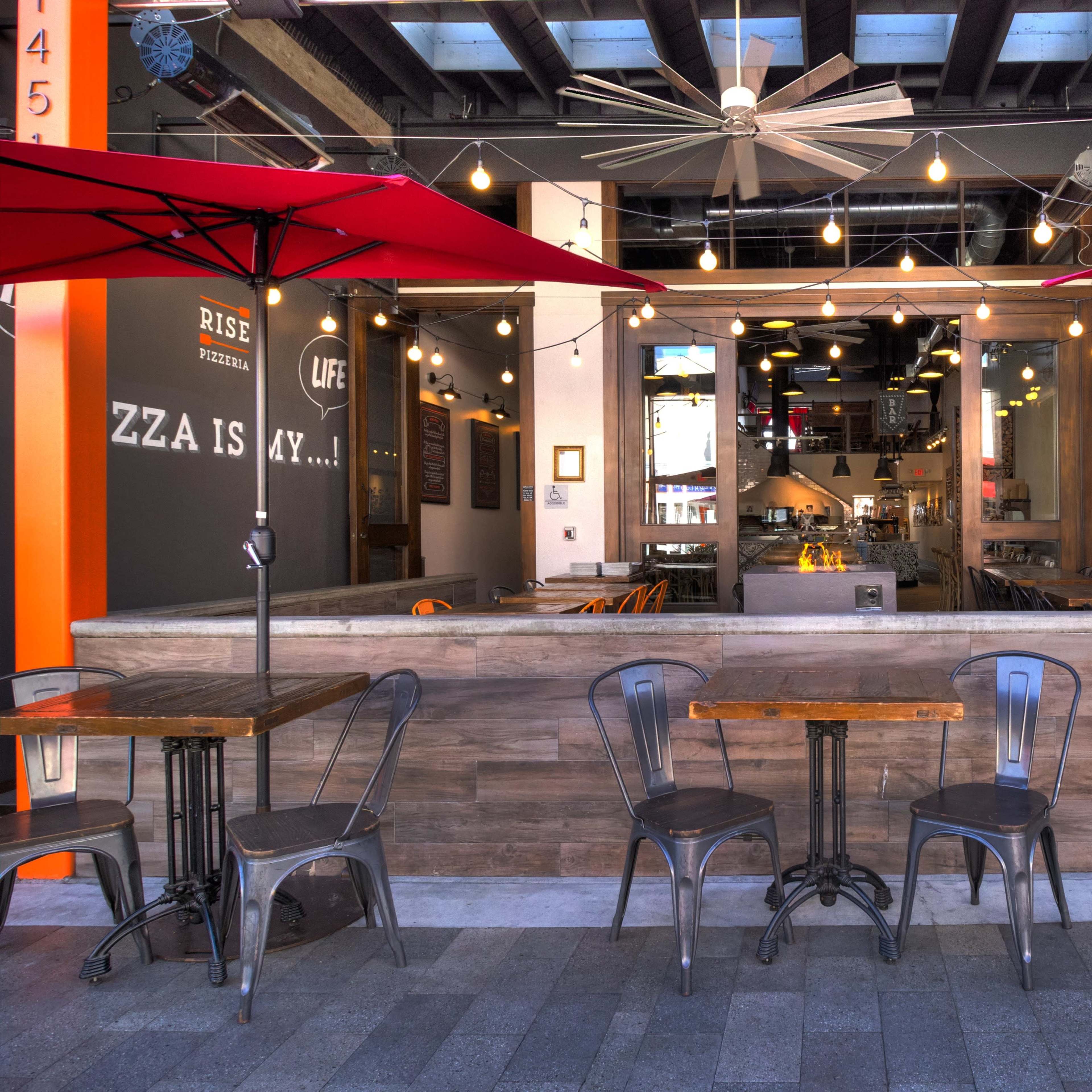 An outdoor seating area of a restaurant with metal tables and chairs beneath red umbrellas, facing an interior space with wooden accents and hanging lights.