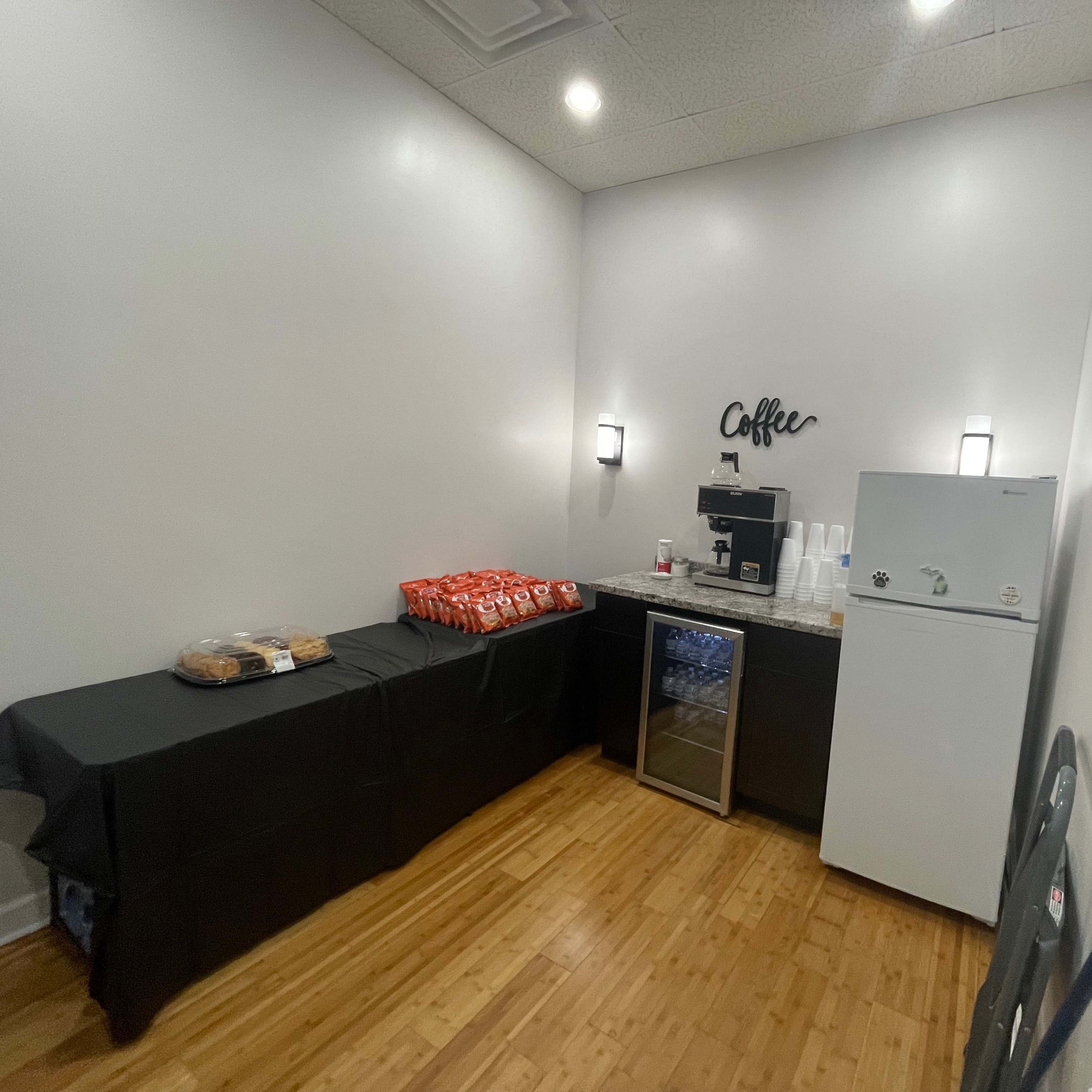 A small break room featuring a coffee station with a coffee maker, a fridge, and a table covered with a black cloth displaying snacks.