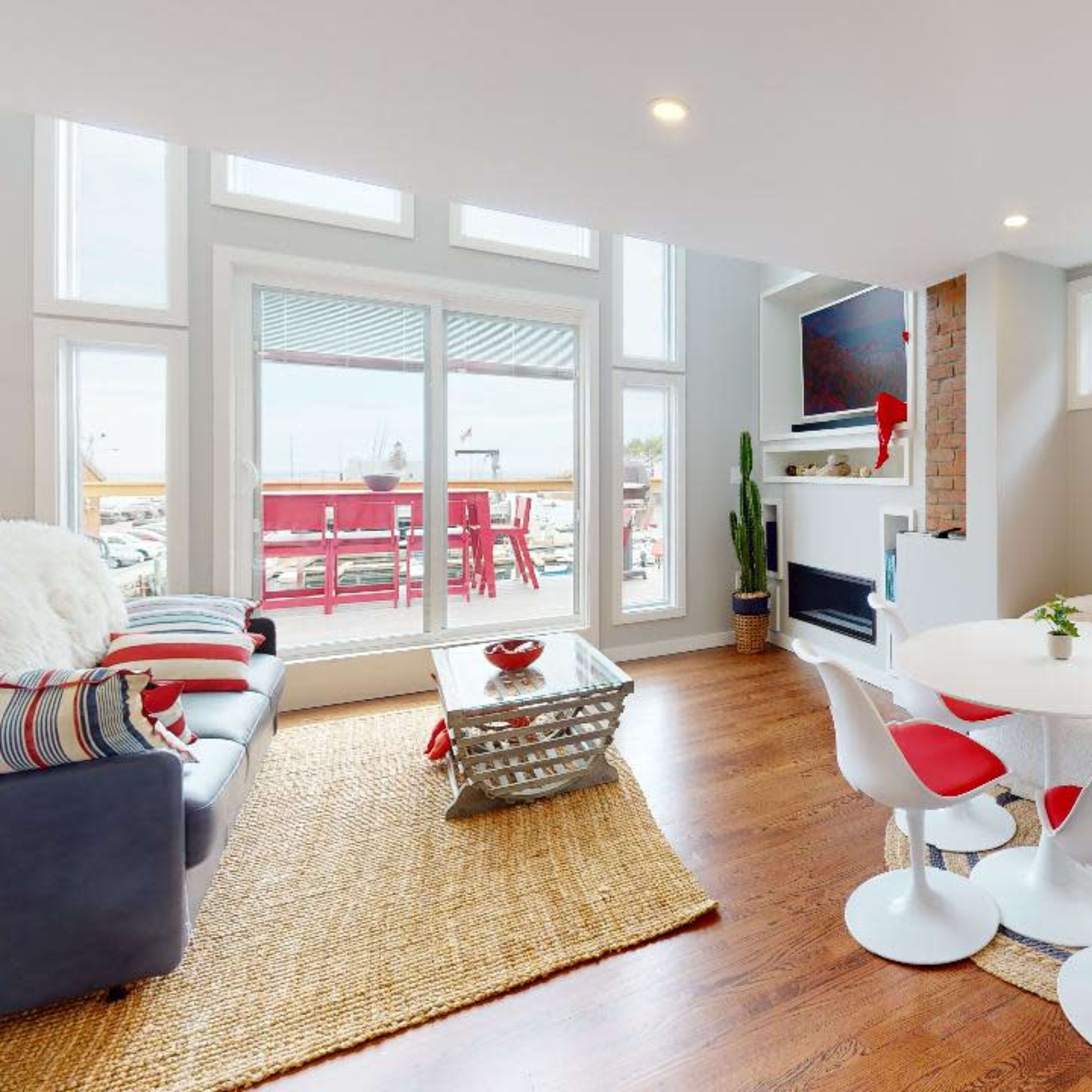 A modern living room features a light-colored sofa, a round dining table with red and white chairs, and large windows overlooking a patio.