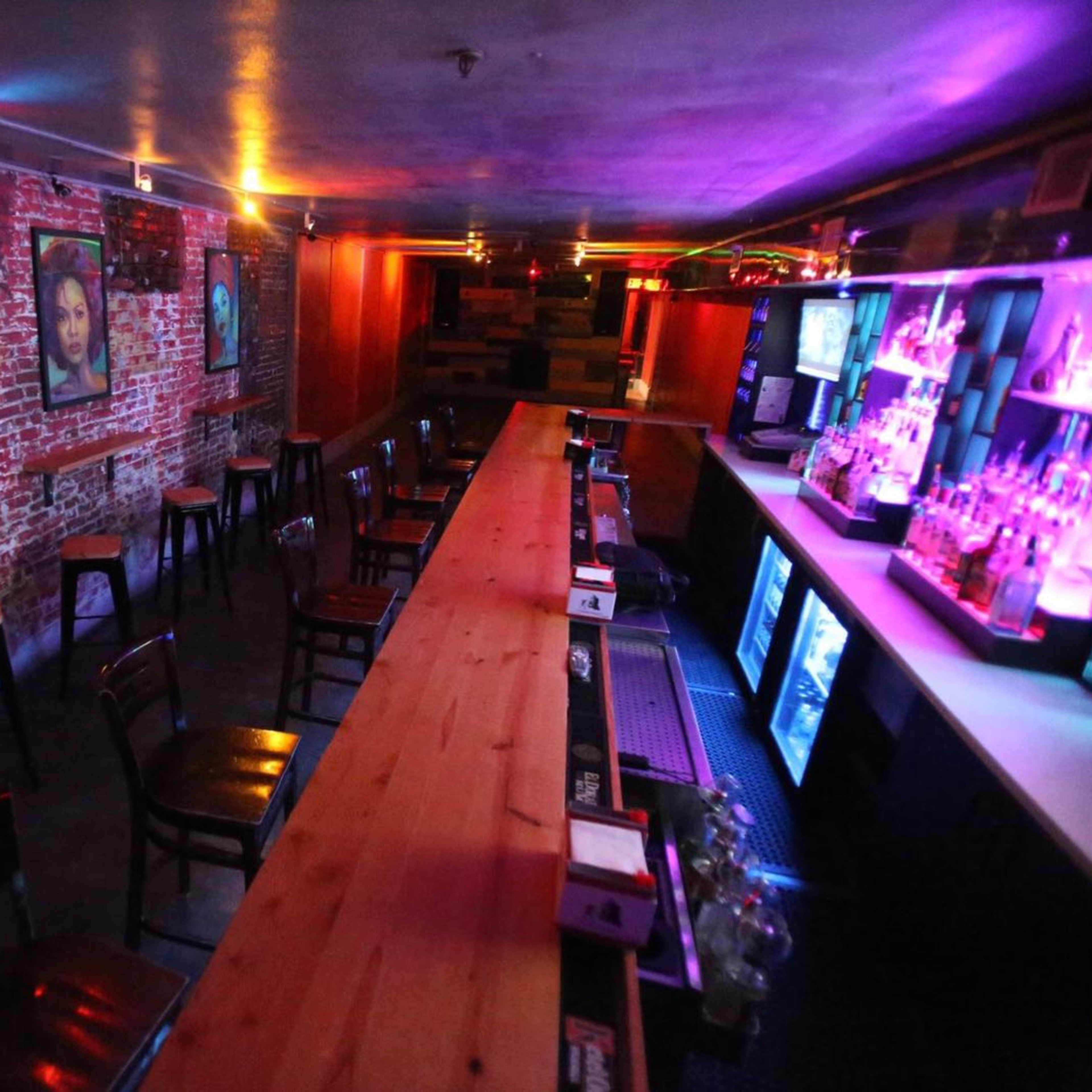 An empty bar area featuring a long wooden counter, high-backed chairs, exposed brick walls with framed artwork, and colorful lighting.