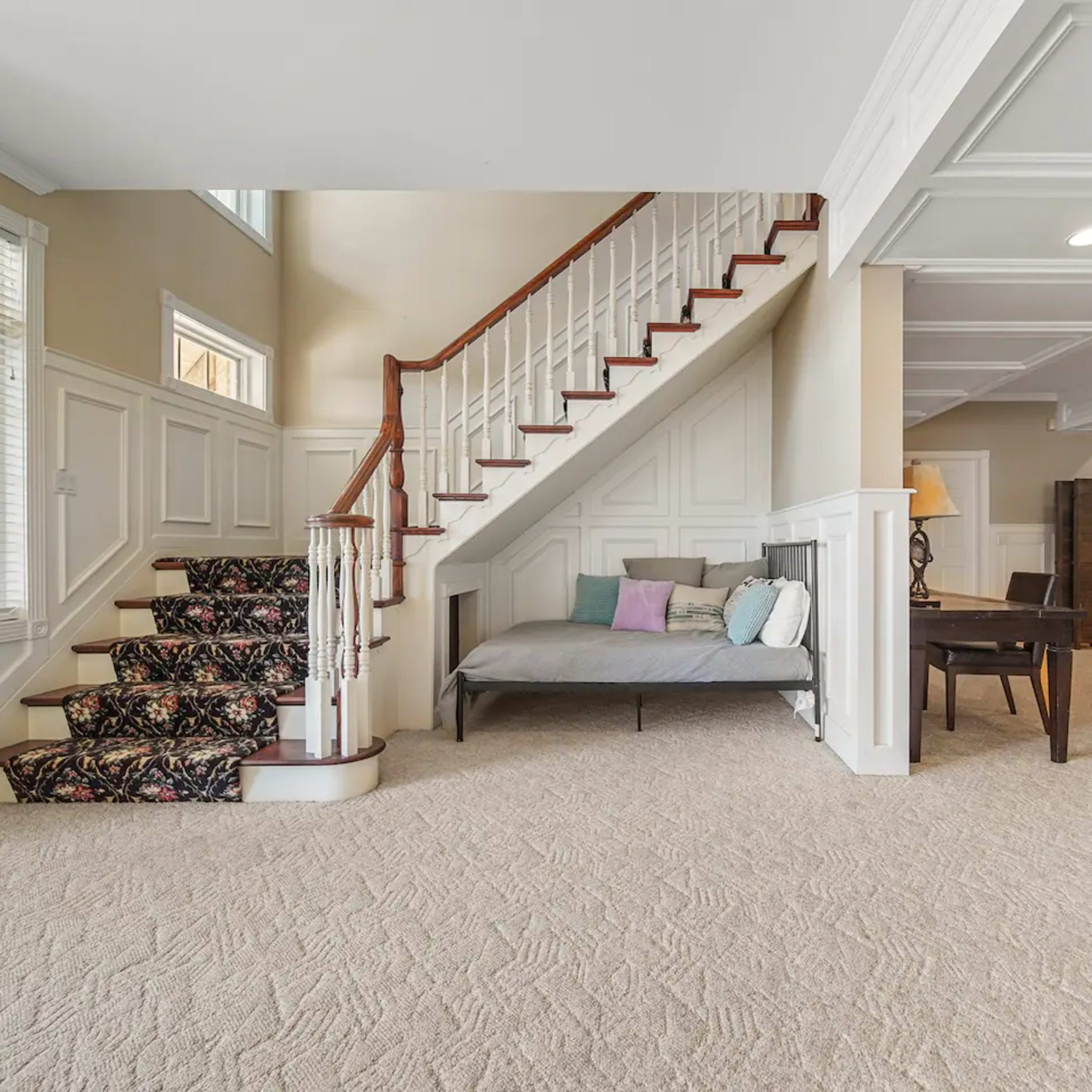 A spacious living area with a staircase, a seating area, a desk, and a bunk bed in a well-lit basement.