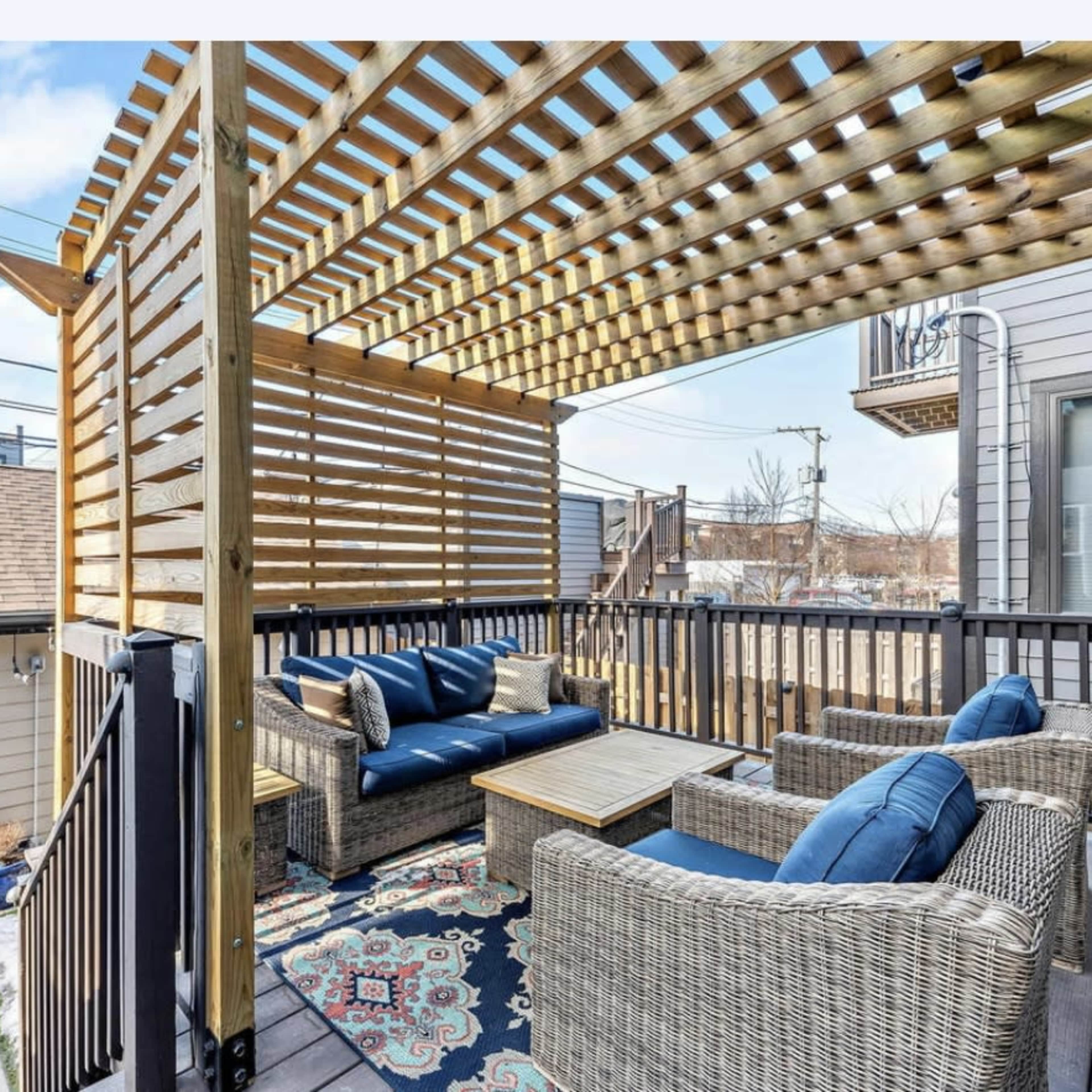 An outdoor seating area with wicker furniture, a wooden pergola overhead, and a patterned rug on a deck.