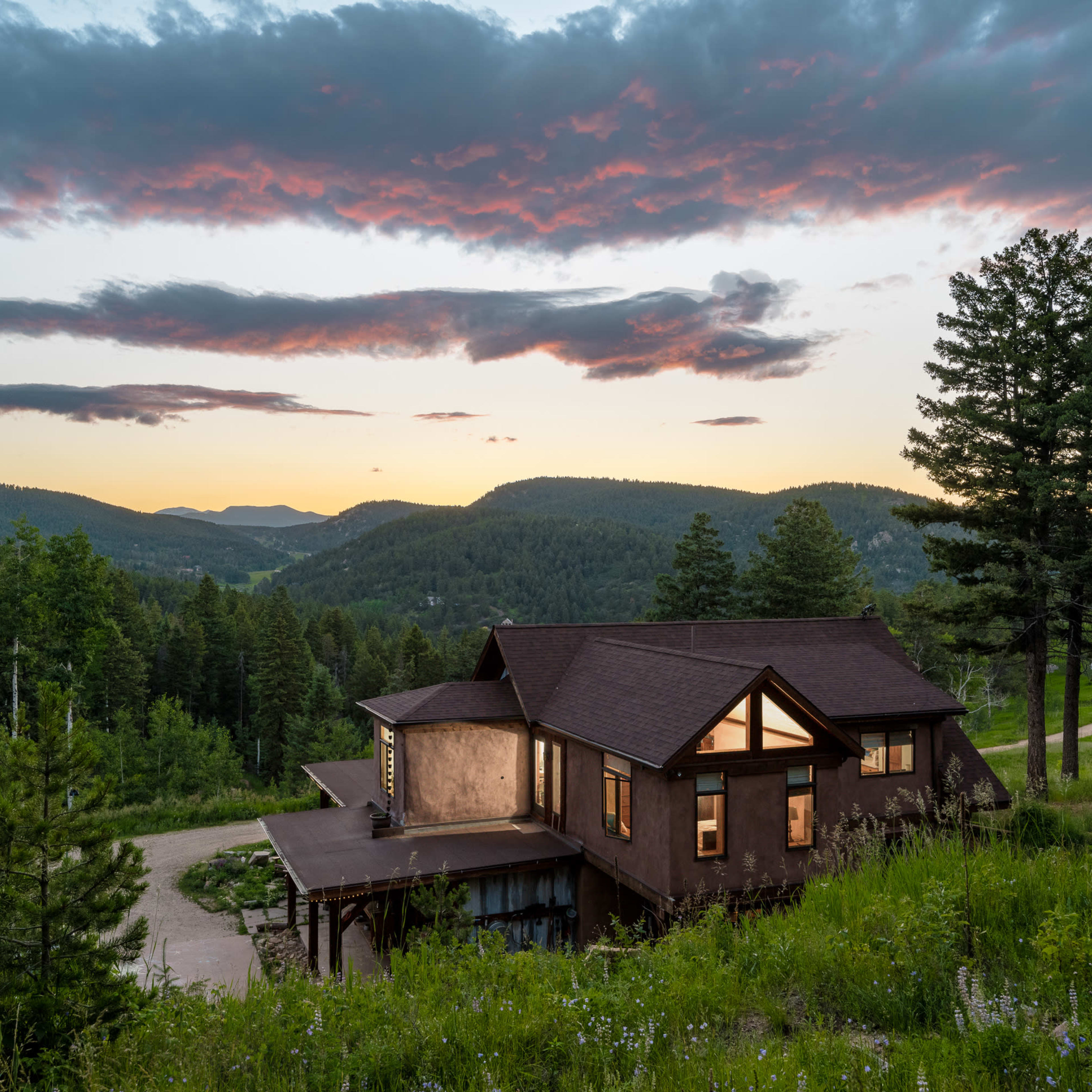 A modern house sits on a hillside, surrounded by trees and rolling mountains under a colorful sunset sky.