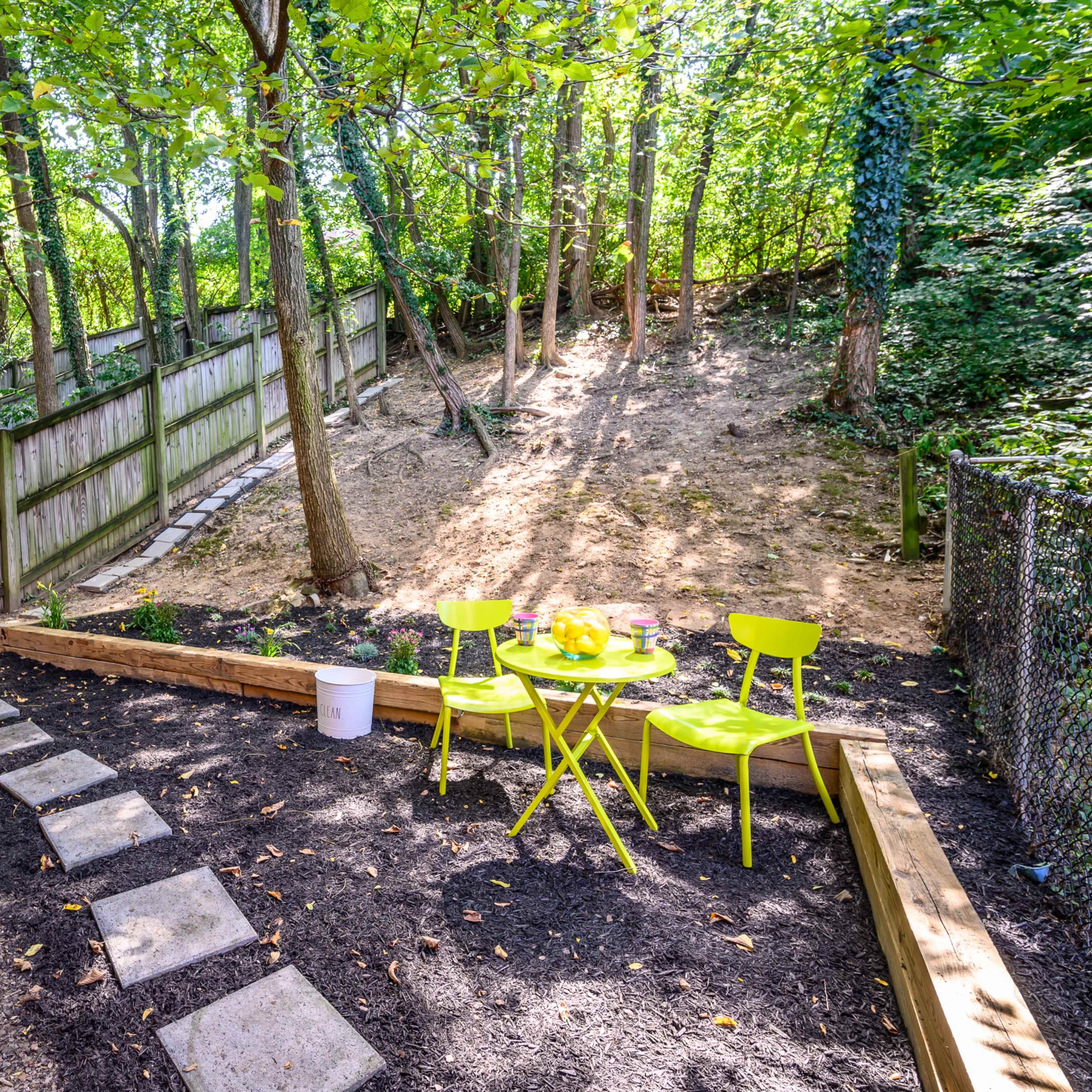 A small, fenced garden area features a wooden seating arrangement with bright chairs and a table, surrounded by trees and a sloped, sandy hillside.