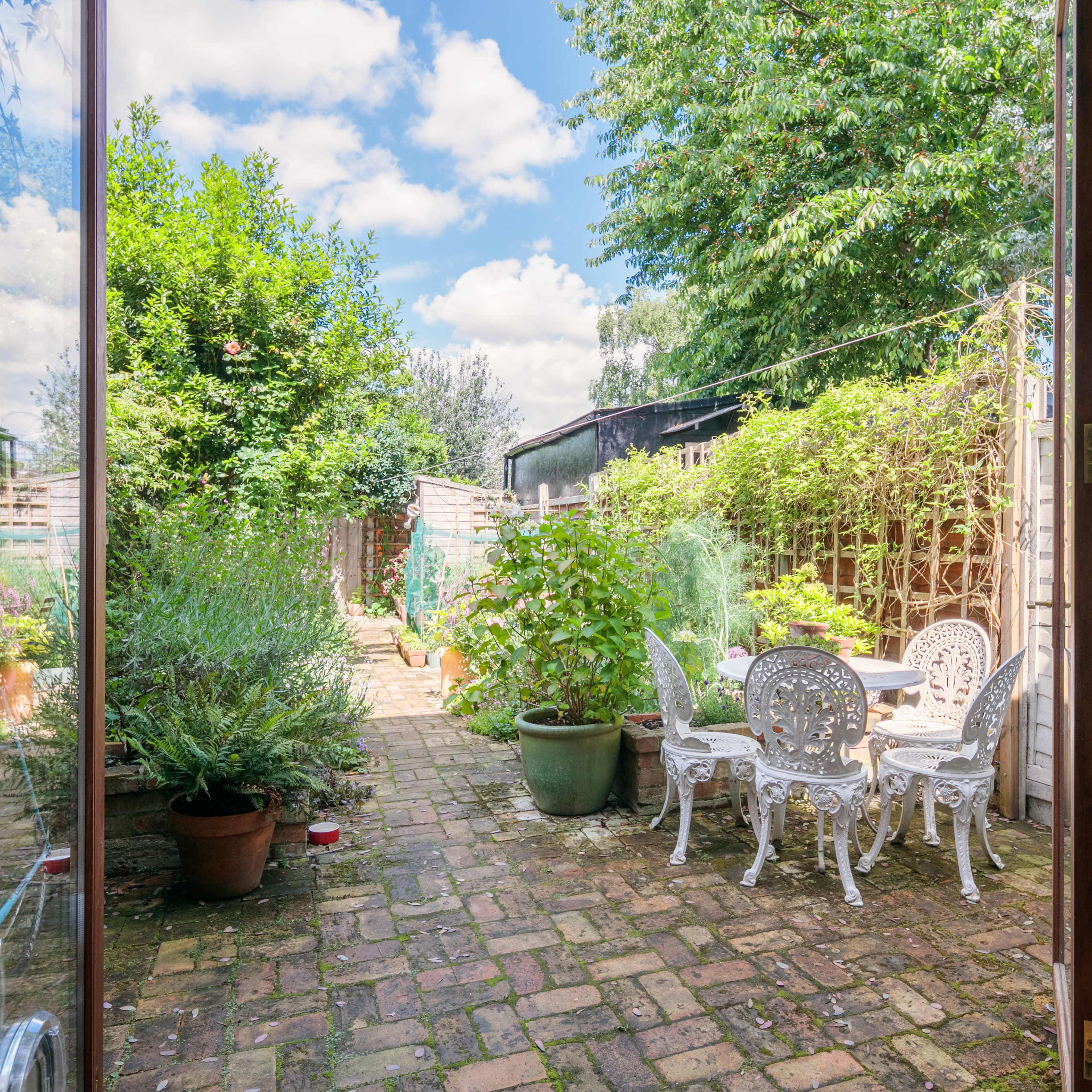 A view of a garden with brick pathways, potted plants, and white outdoor furniture, as seen through open glass doors.