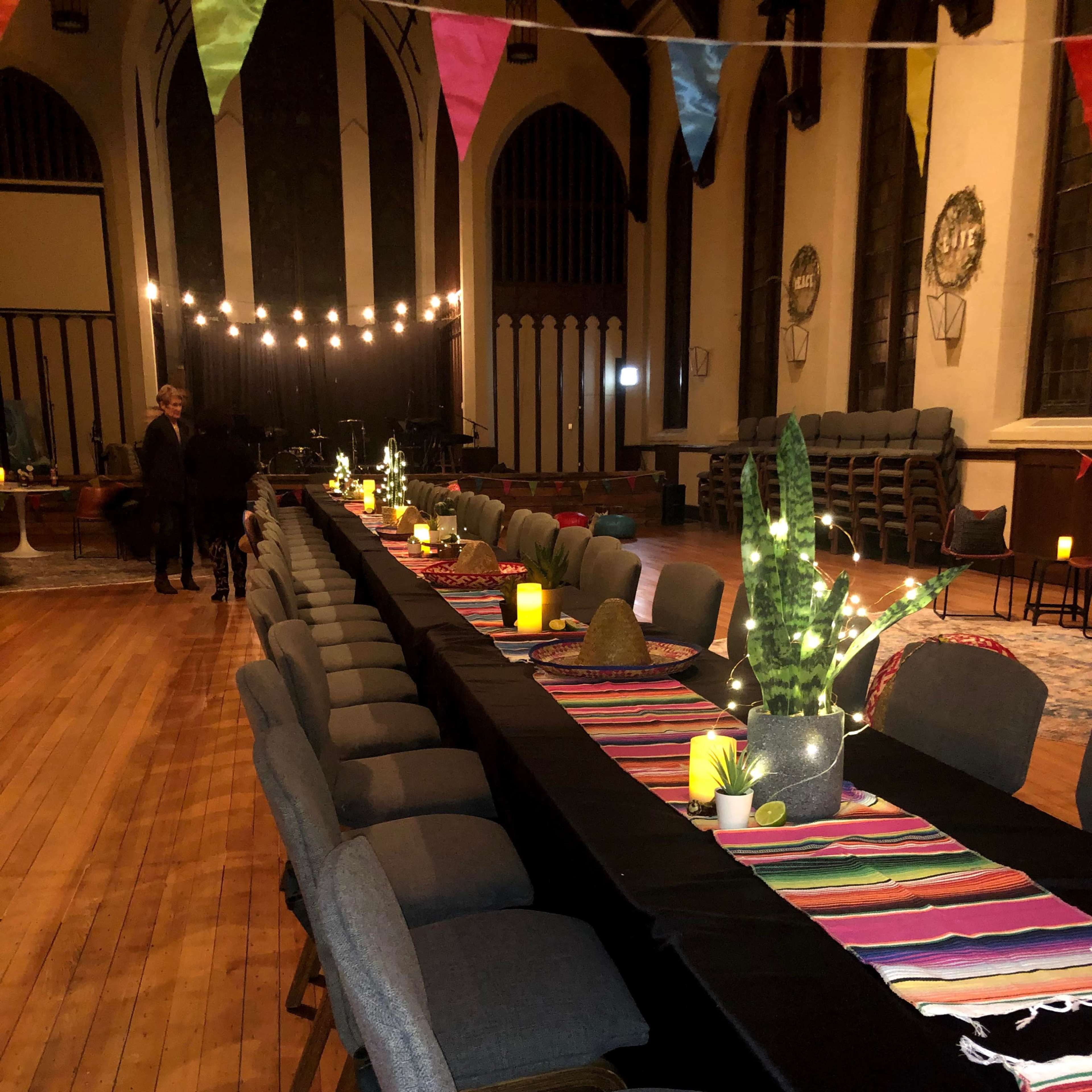 A long table with gray chairs is set in a large room adorned with colorful banners and string lights, featuring a striped table runner and decorative plants.