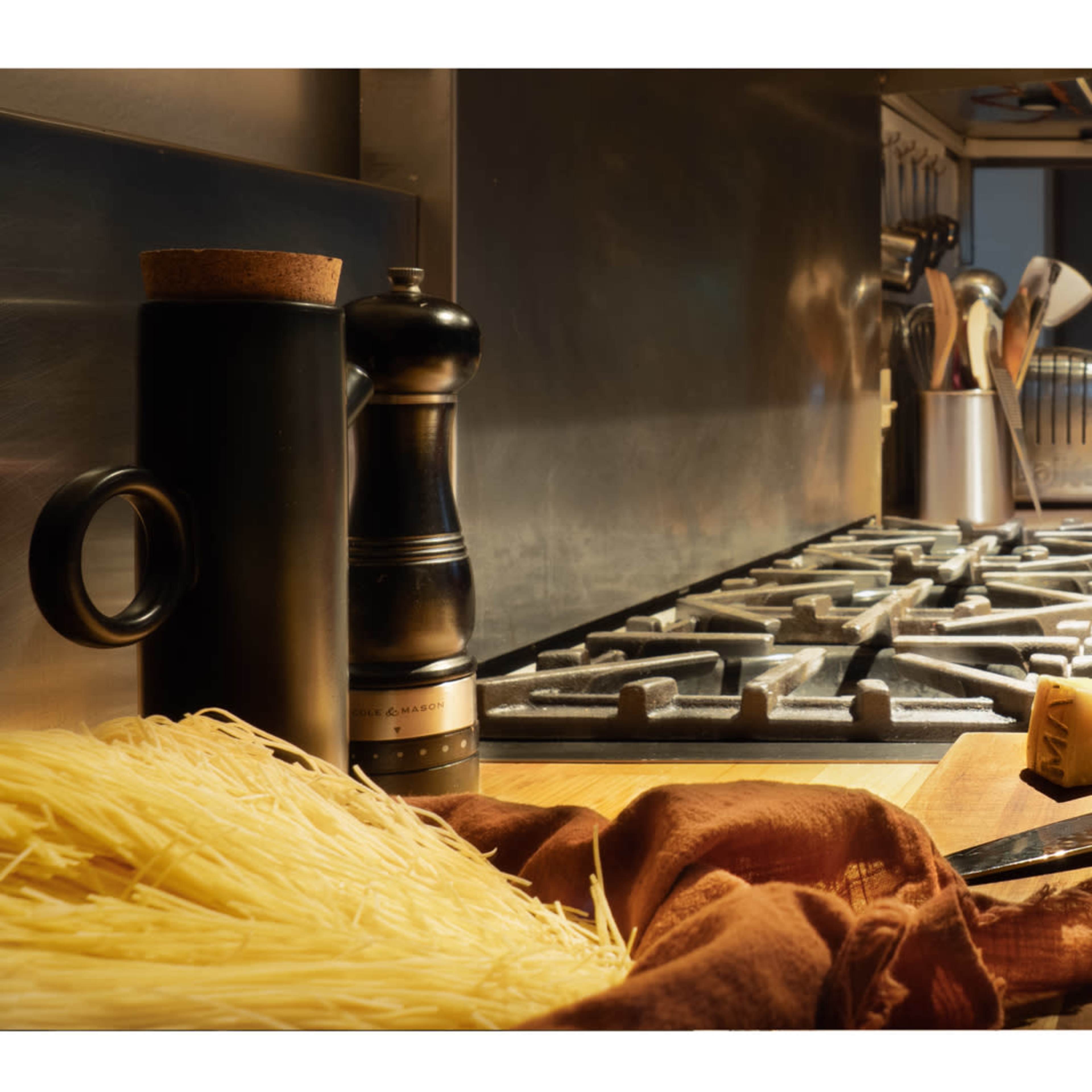 A kitchen scene features a red pot on a stove, surrounded by uncooked pasta, a knife, a lemon, cheese, and a pepper mill on a wooden cutting board.