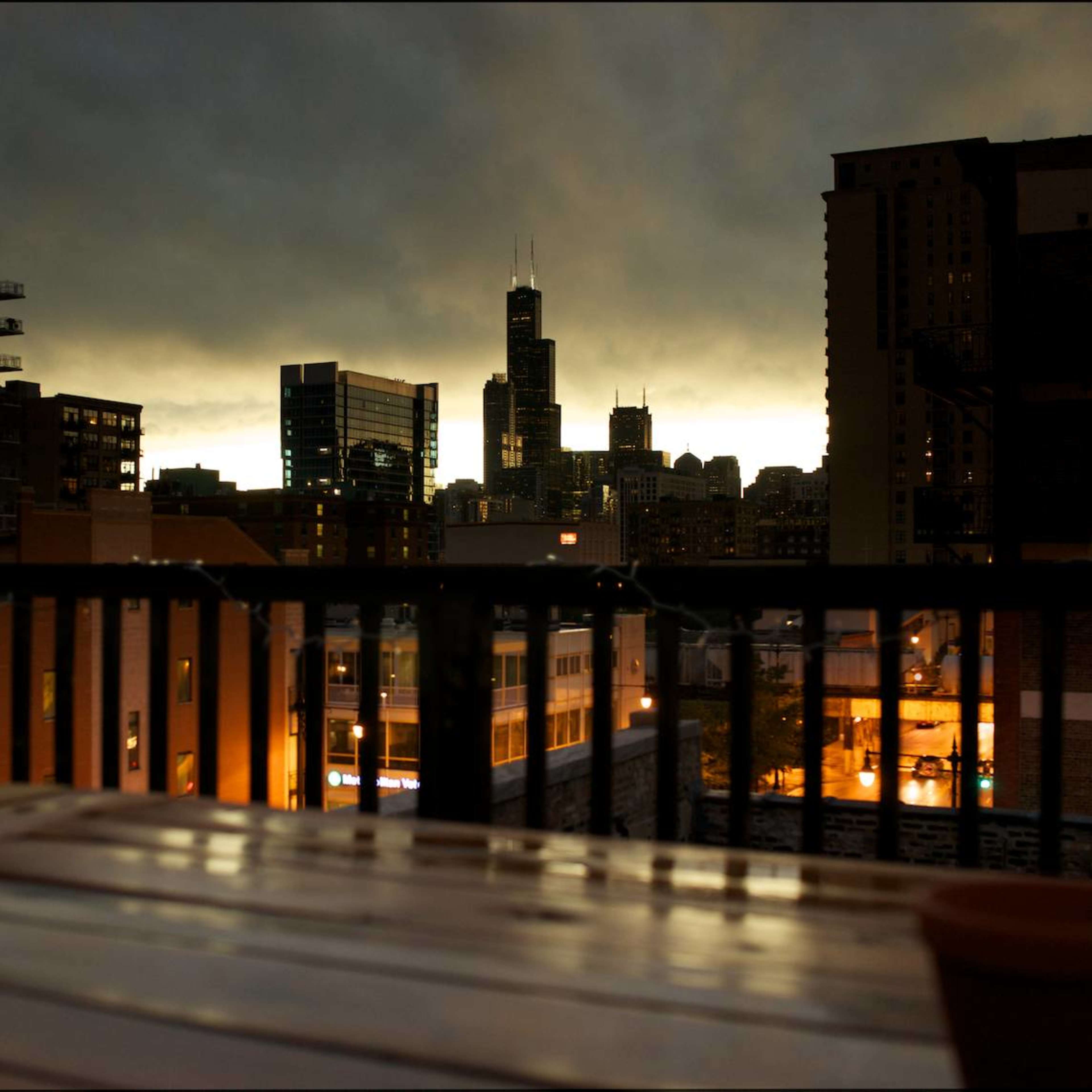 A city skyline at dusk, with buildings silhouetted against a cloudy sky and lights starting to illuminate in the neighborhoods below.