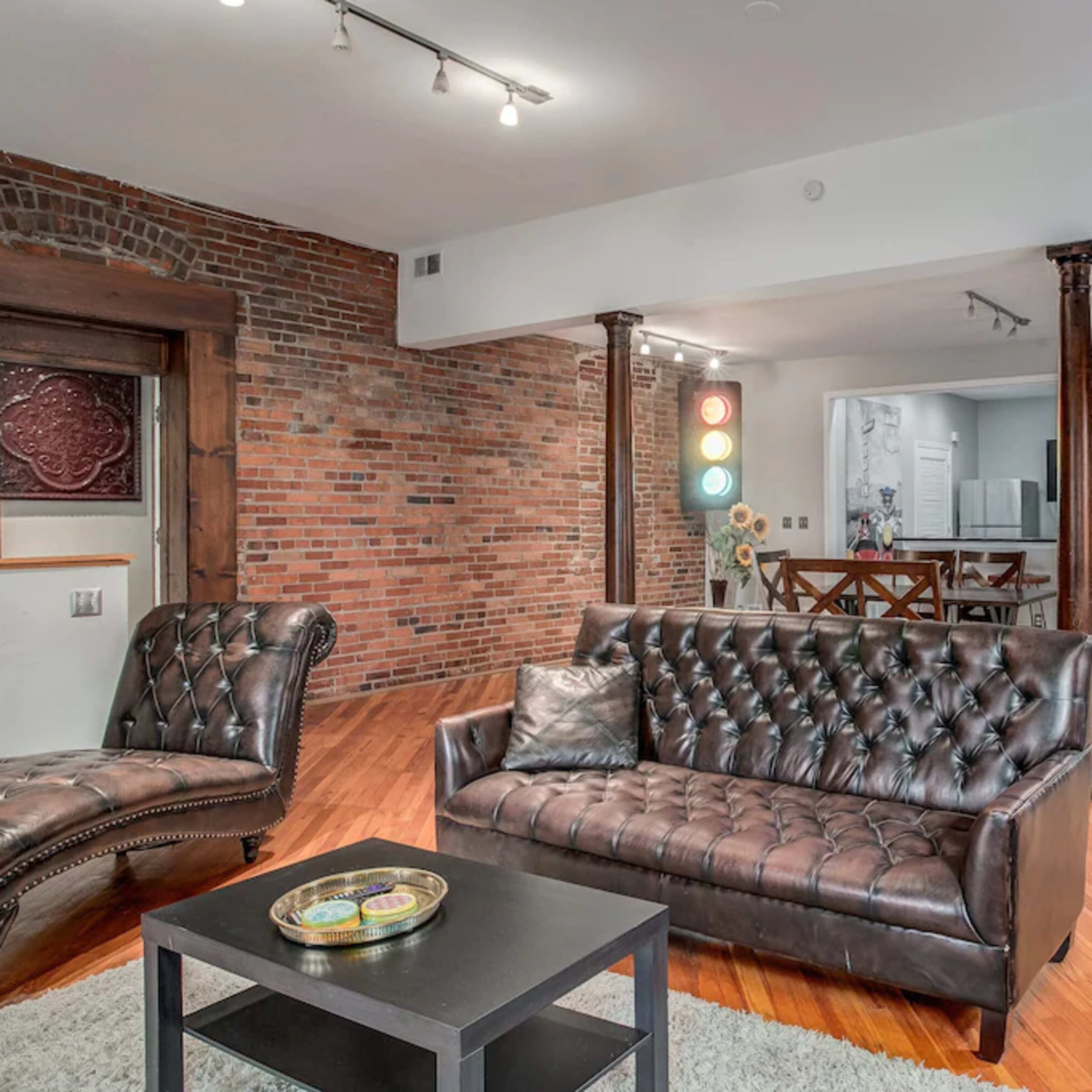 A spacious living area featuring a brown leather couch, a matching chair, hardwood flooring, exposed brick walls, and a glimpse of a kitchen in the background.