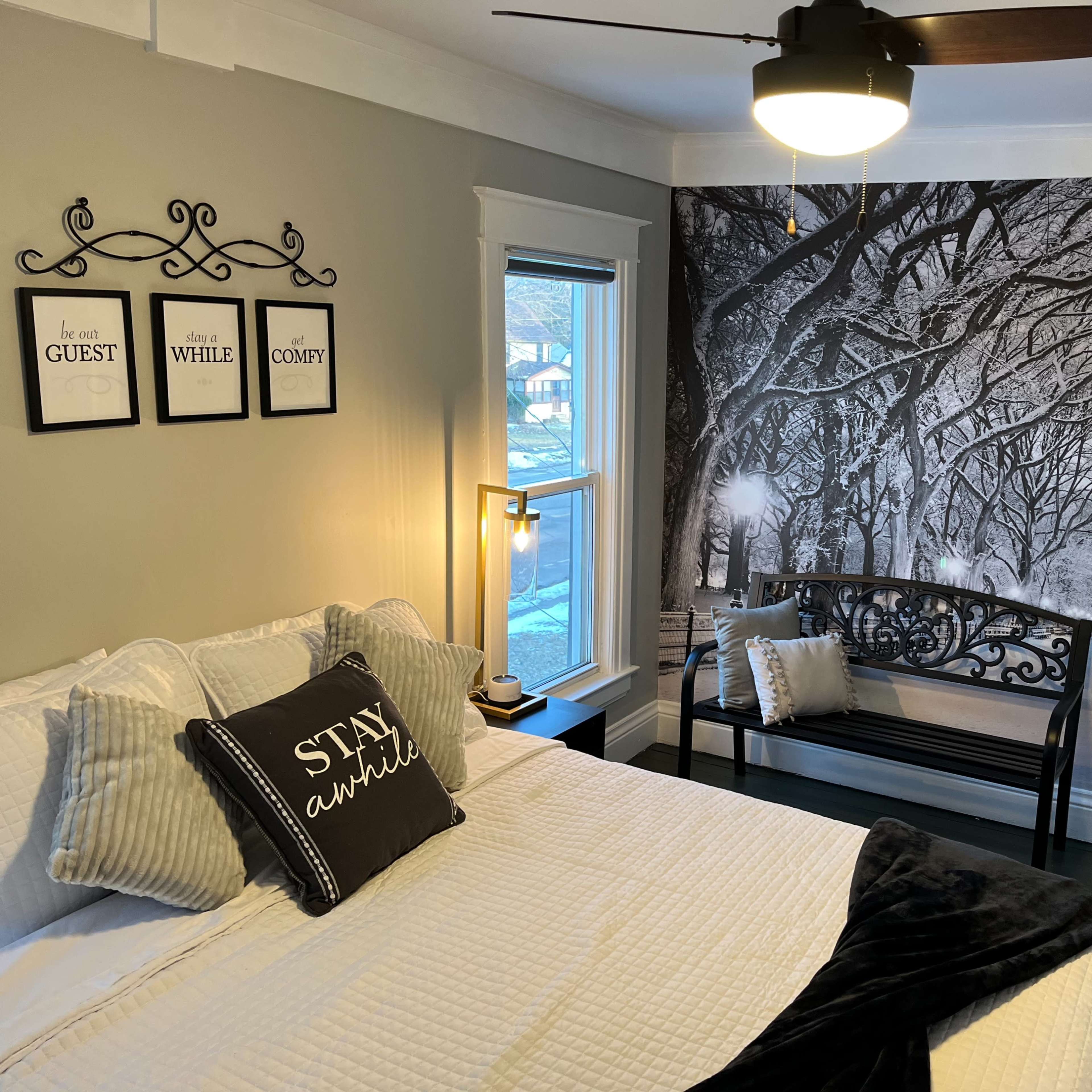 A cozy bedroom with a bed, a decorative bench, and a large black-and-white mural of a tree-lined street on one wall.