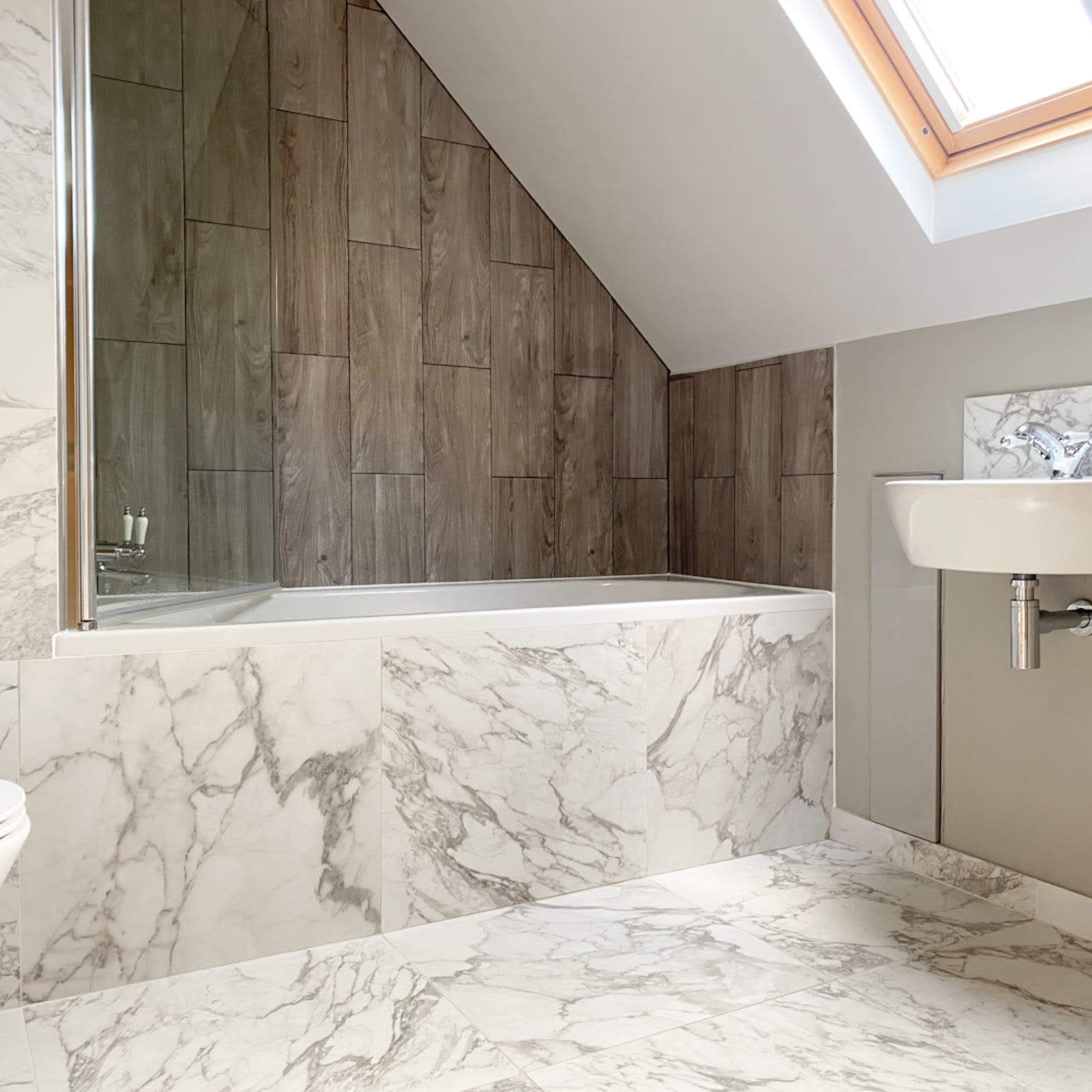 A bathroom with a marble floor, a bathtub, and a wall with wooden tiles under a slanted ceiling with a skylight.