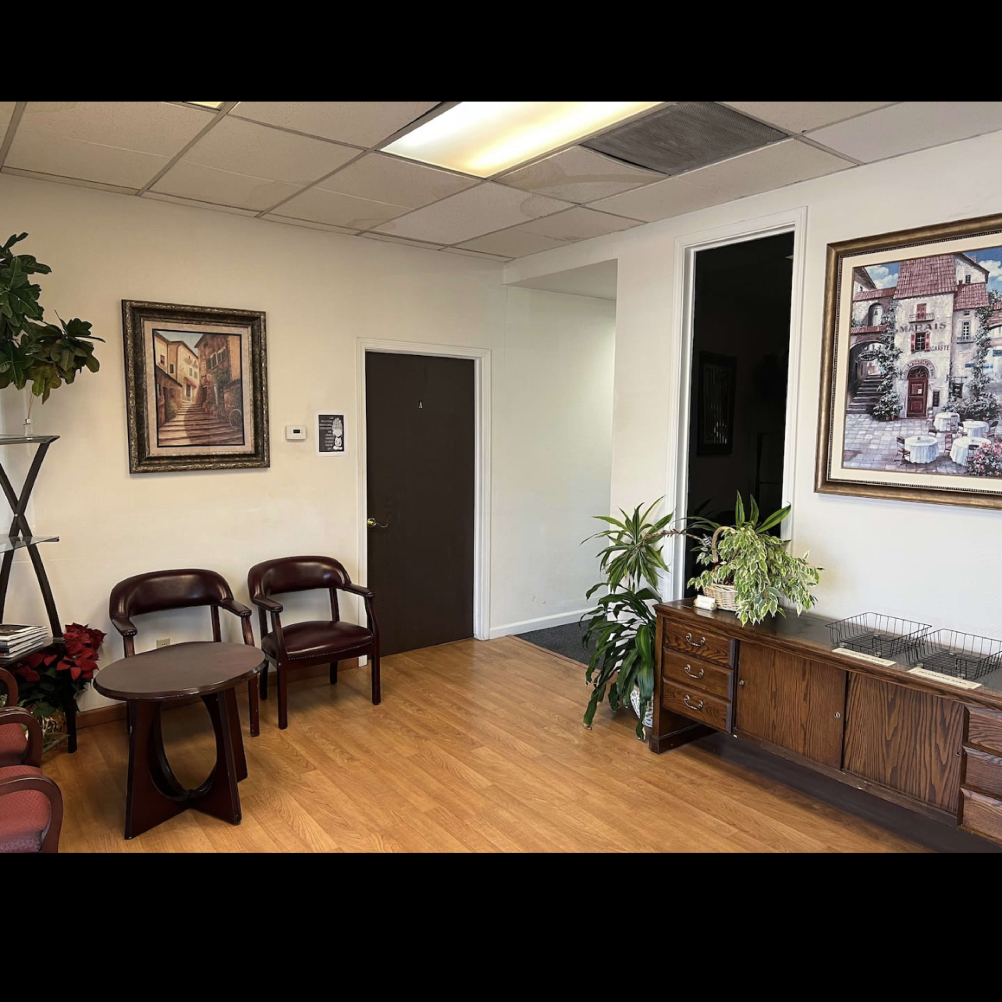 A reception area with two wooden chairs, a small table, and potted plants, alongside a painting on the wall and a cupboard.